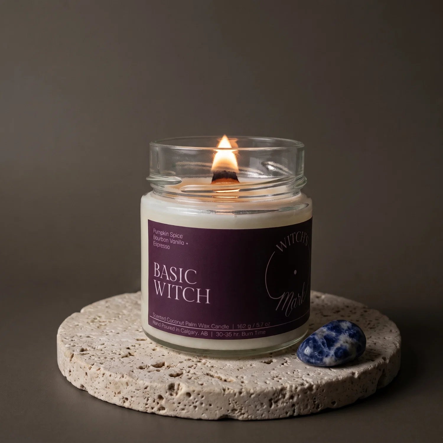 A lit Basic Witch crystal candle sits on a porous stone coaster with a sodalite crystal next to it.