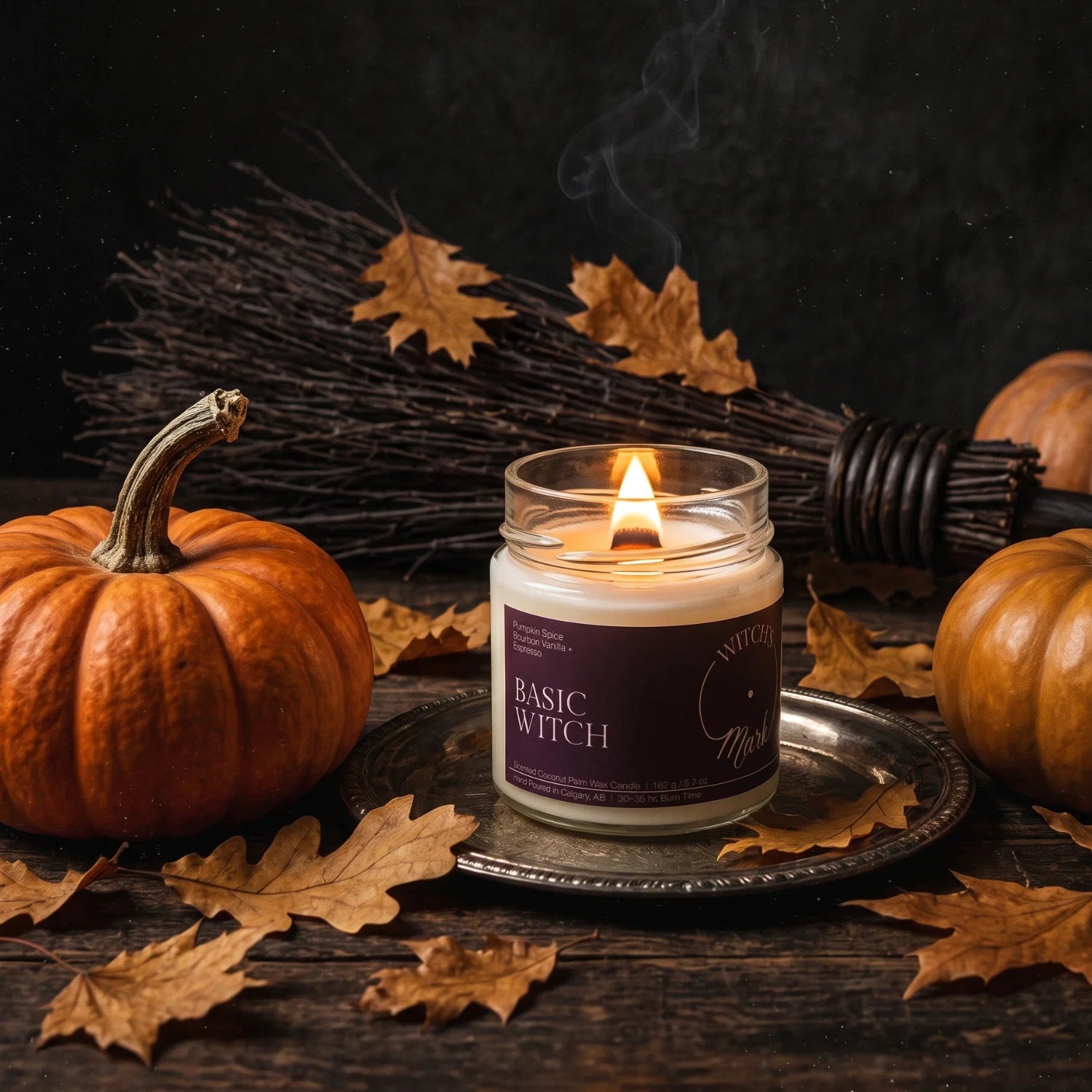 A Basic Witch candle sitting on a brass plate surrounded by an orange pumpkin, a witch's besom and autumn leaves.