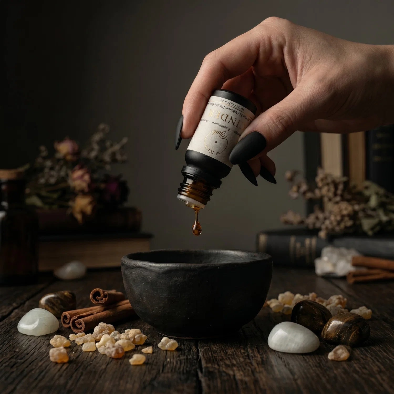 Hand pouring oil from a bottle labeled 'CINDER' into a dark bowl on a wooden surface with frankincense tears, cinnamon sticks and stones.