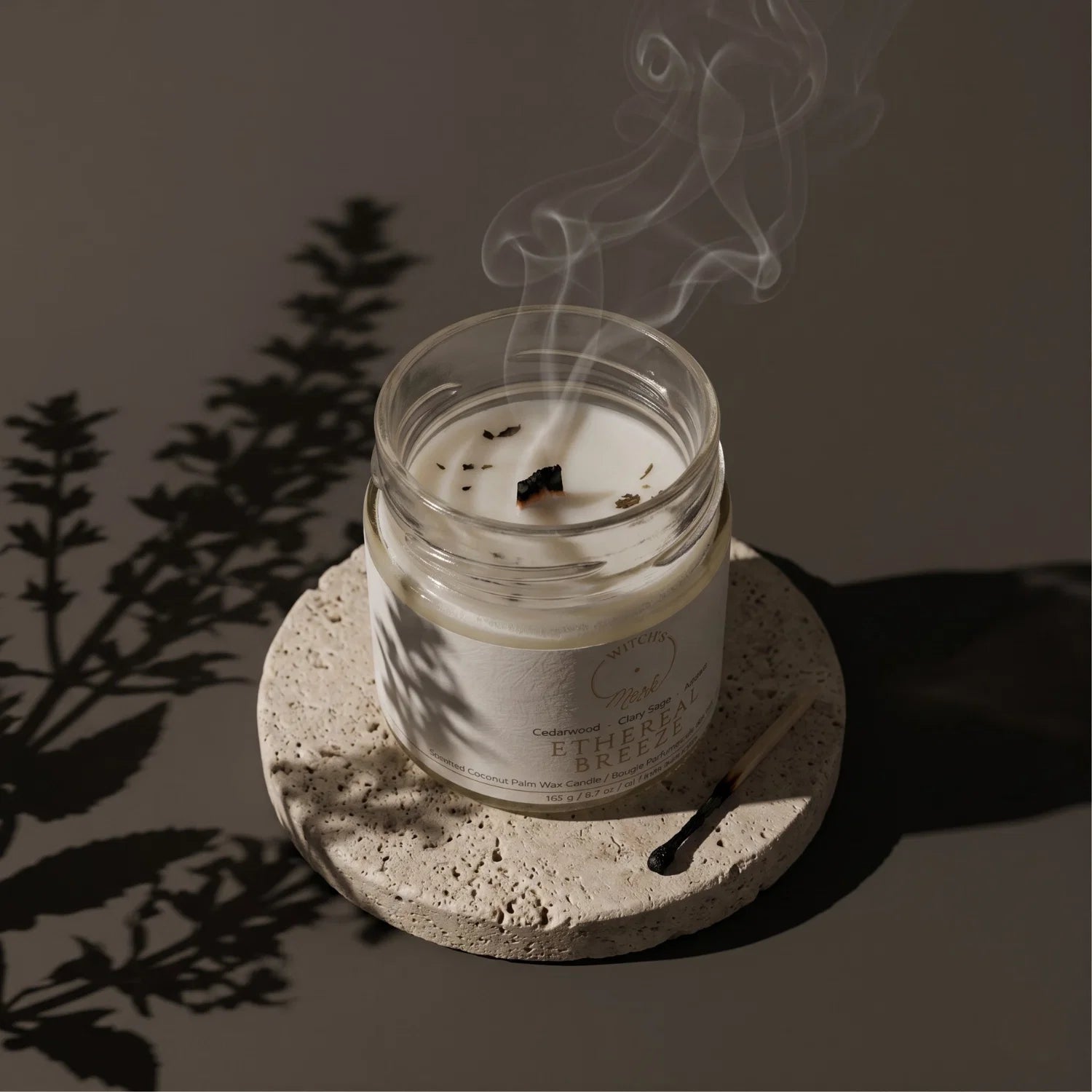 Ethereal Breeze crystal candle extinguished with smoke dancing up from the unlit wick. The candle is on a coaster and next to the coaster is a burnt match.