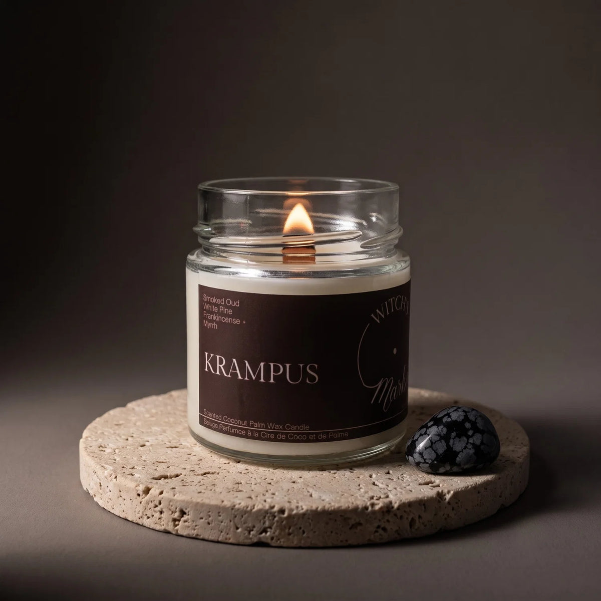 A lit Witch's Mark scented candle in a clear glass jar sits on a round stone coaster, with a dark red label reading 'KRAMPUS' and a snowflake obsidian crystal beside it against a dark gray background.