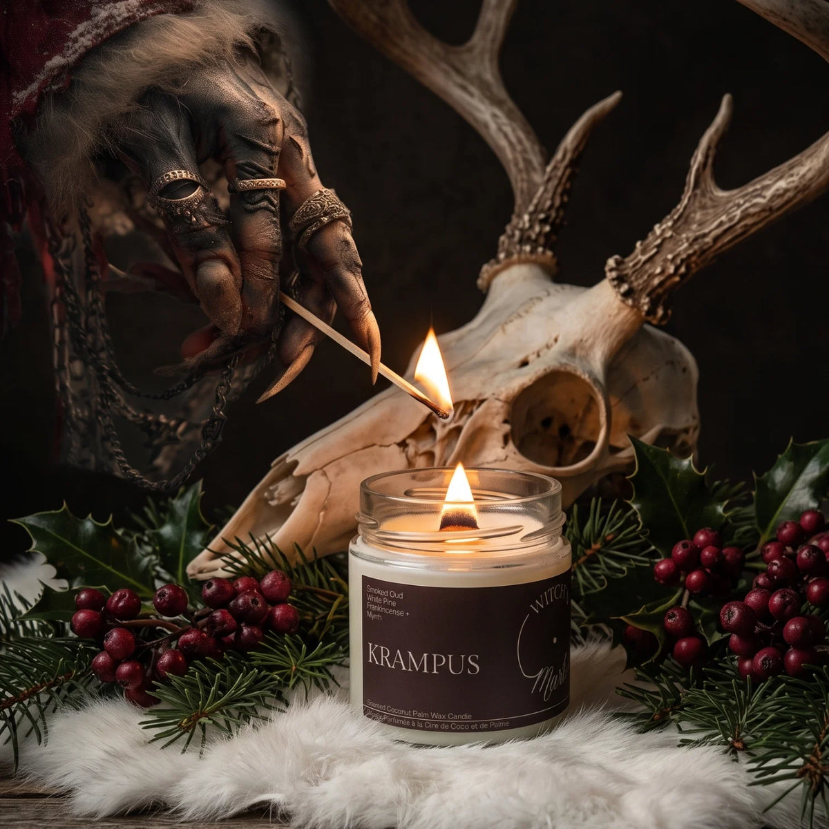A lit candle in a glass jar labeled 'KRAMPUS' sits on a white faux-fur surface, surrounded by holly leaves, red berries, pine branches, a deer skull, antlers, and a dark clawed hand holding a match. The candle is the clear main product, with a moody rustic holiday setting.