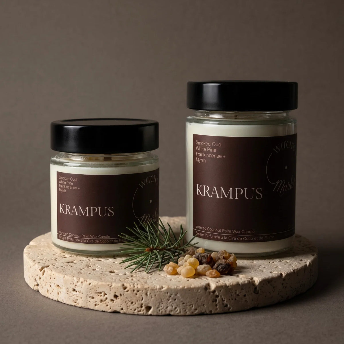 Two Witch's Mark scented wax candles in clear glass jars with black lids and dark red labels reading KRAMPUS, styled on a round stone slab with a small pine sprig and frankincense tears against a taupe background.