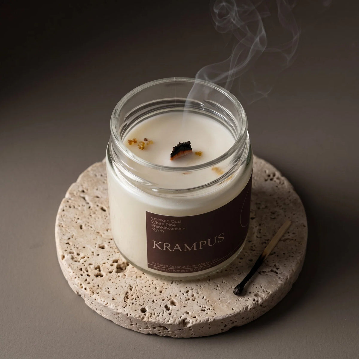 A lit Witch's Mark scented candle in a clear glass jar sits on a round stone coaster, with smoke rising from the surface. The label reads KRAMPUS, and a small match lies beside the jar on a dark taupe background.
