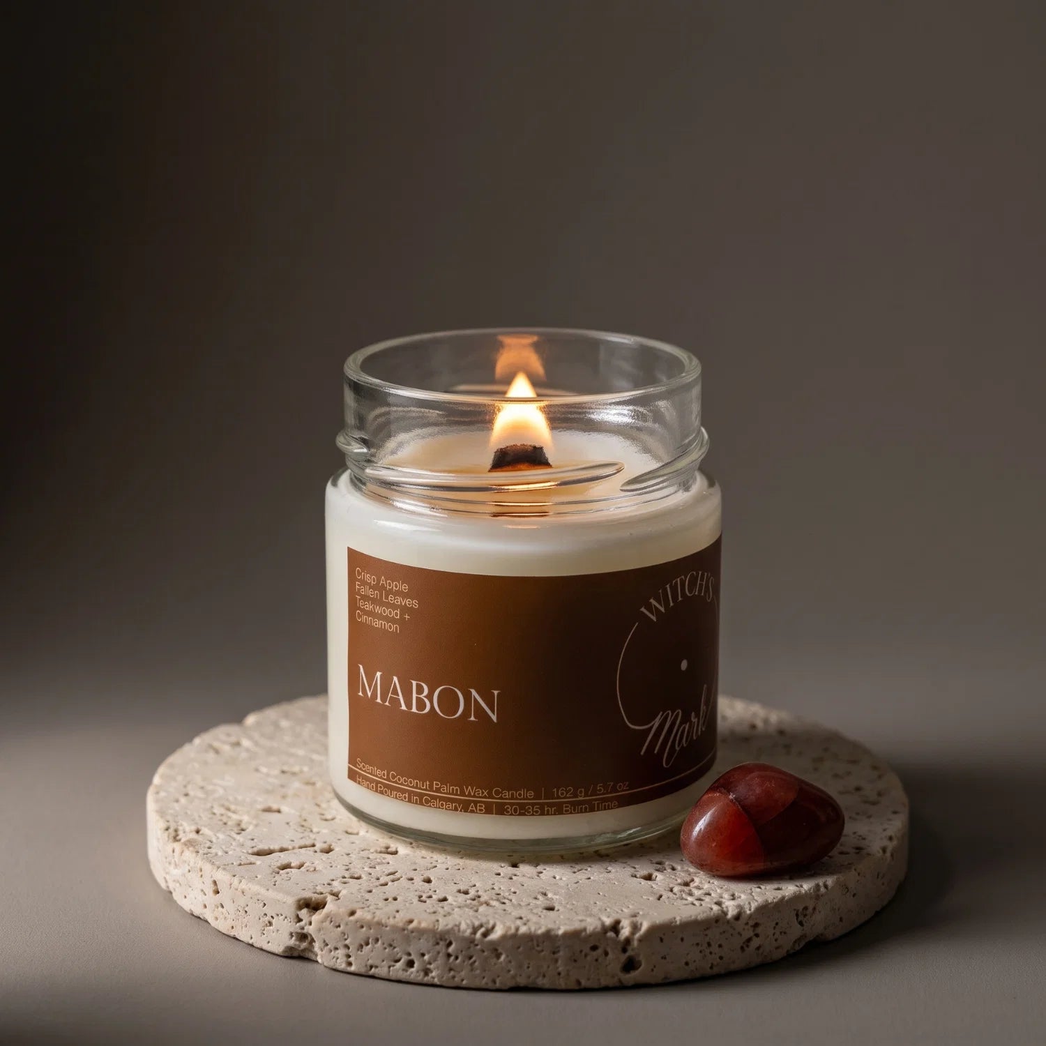 A Mabon crystal candle sits on a porous stone coaster with a red aventurine crystal beside it.