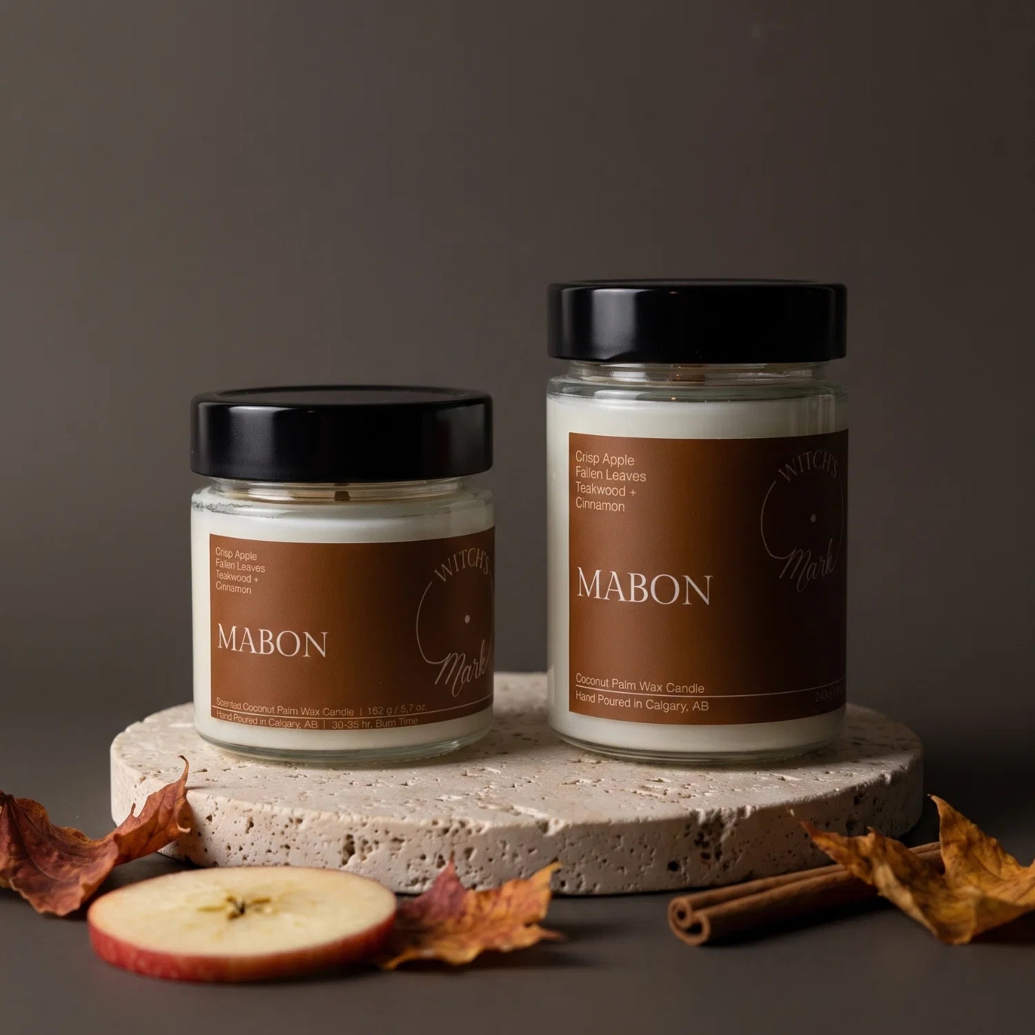 Two Mabon scented candles in clear glass jars with black lids and brown labels, displayed on a round stone pedestal with autumn leaves, cinnamon sticks, and an apple slice against a dark gray background.