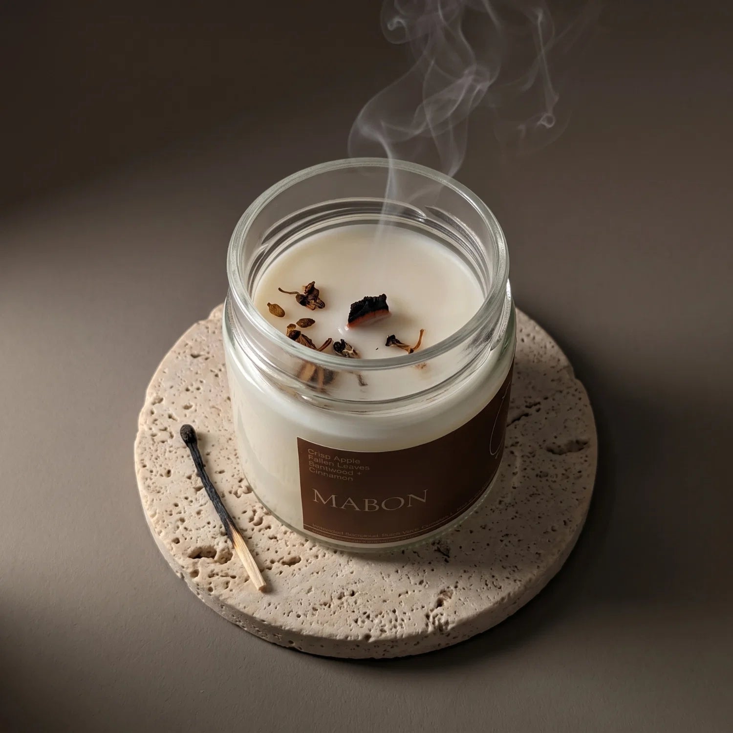 A top view of a candle called "Mabon" where the wick has recently been extinguished with curls of white smoke rising. The candle sits on a porous stone coaster and next to the candle is a match.