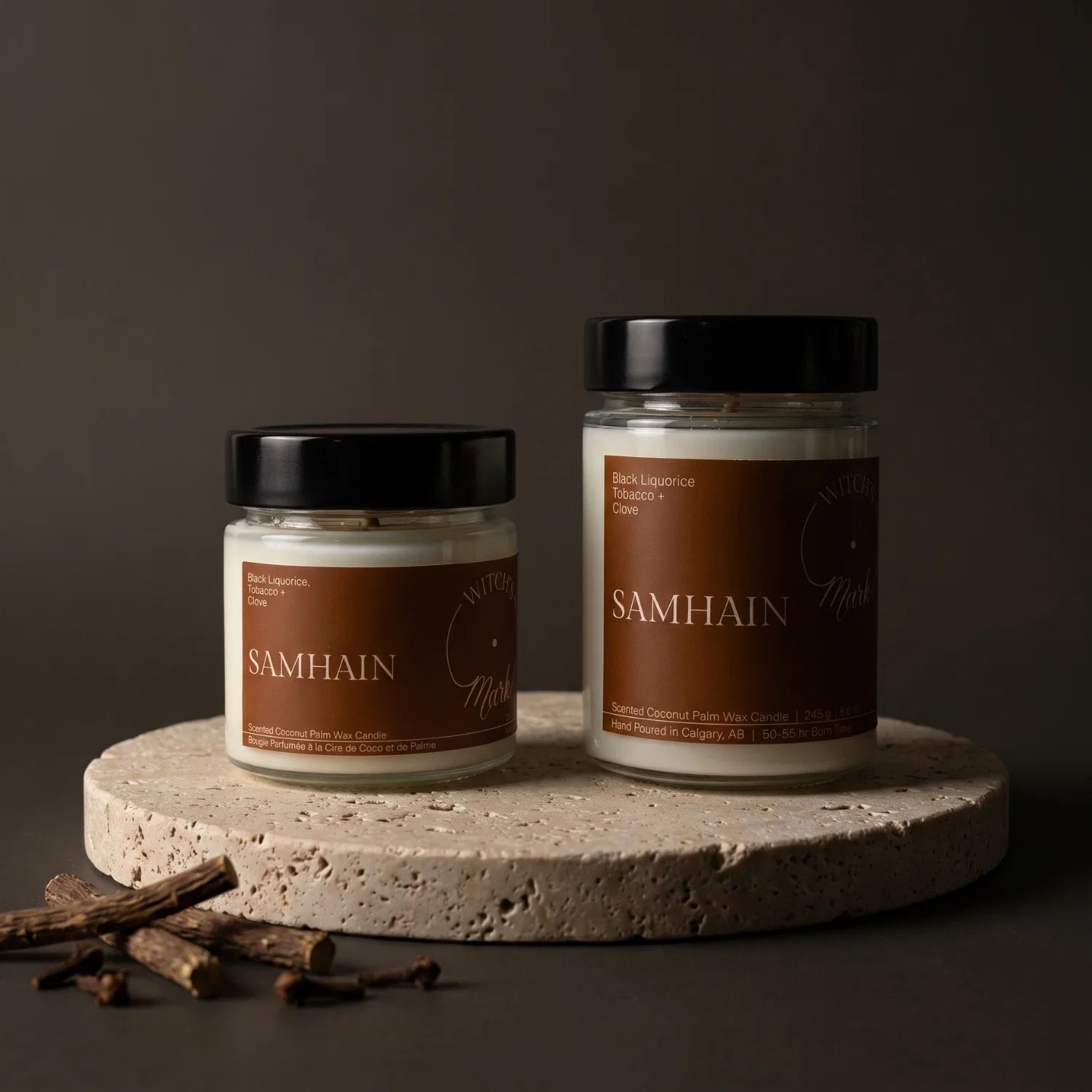 Two glass jar candles with black lids and brown labels reading SAMHAIN, displayed on a round stone platter against a dark brown background, with liquorice root and clove in the foreground.