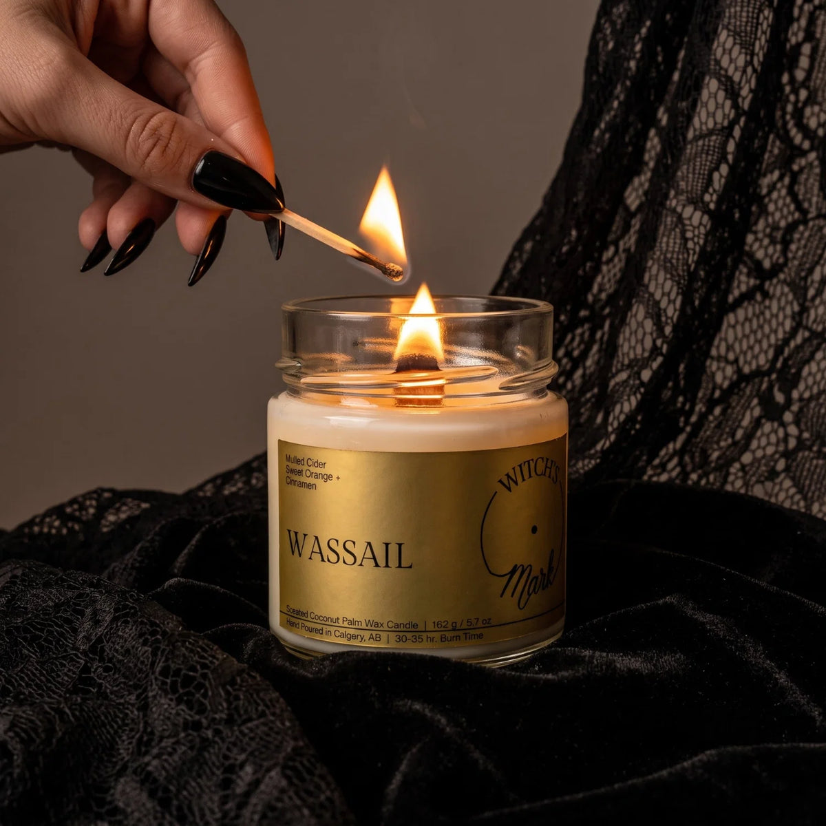 A glass jar candle labeled 'WASSAIL' with a gold label and white wax is lit with a wooden match held by a hand with black manicured nails, set against a dark lace fabric background.