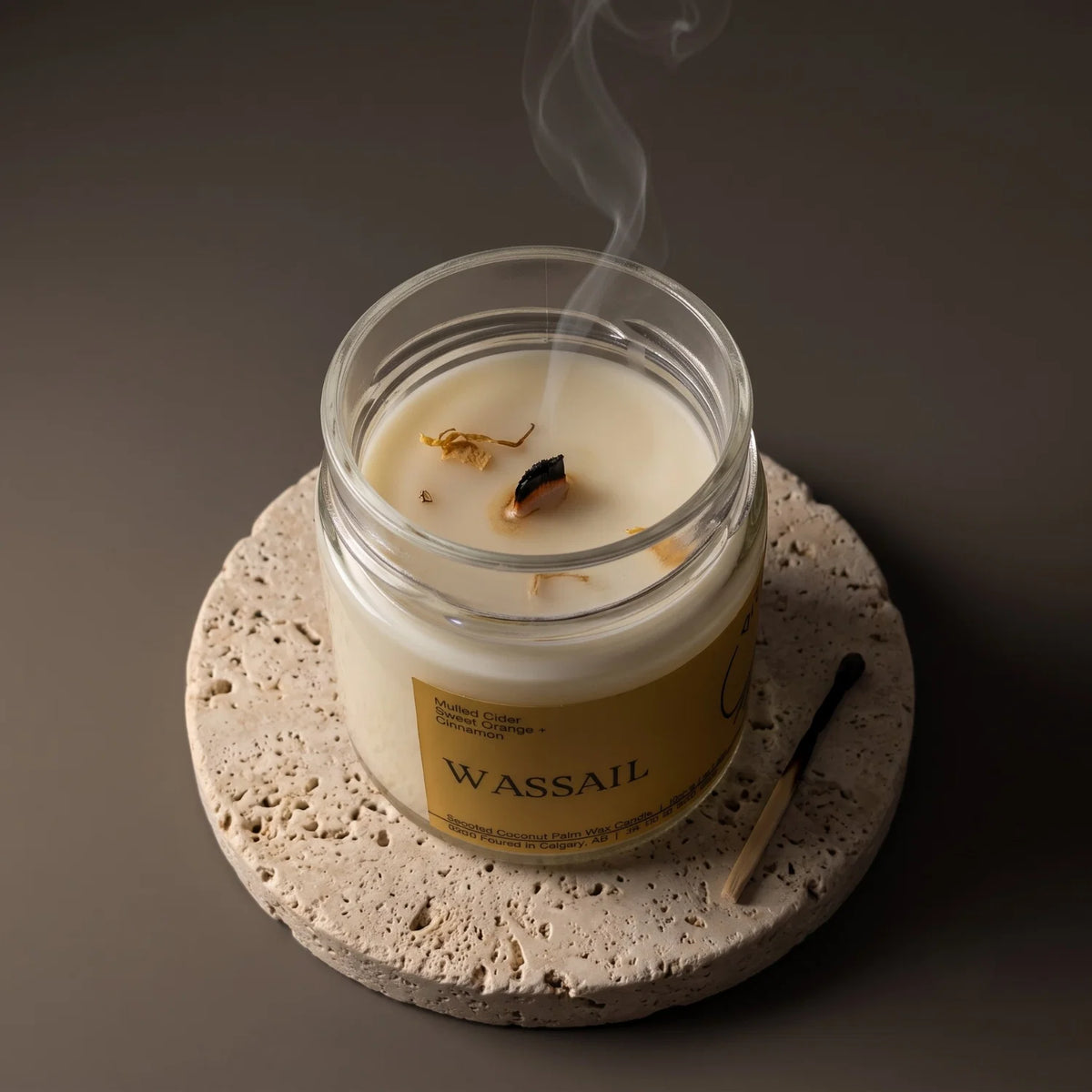 A glass jar candle with creamy off-white wax sits on a round stone coaster, with a match beside it and a thin wisp of smoke rising from the wax. The label reads WASSAIL.