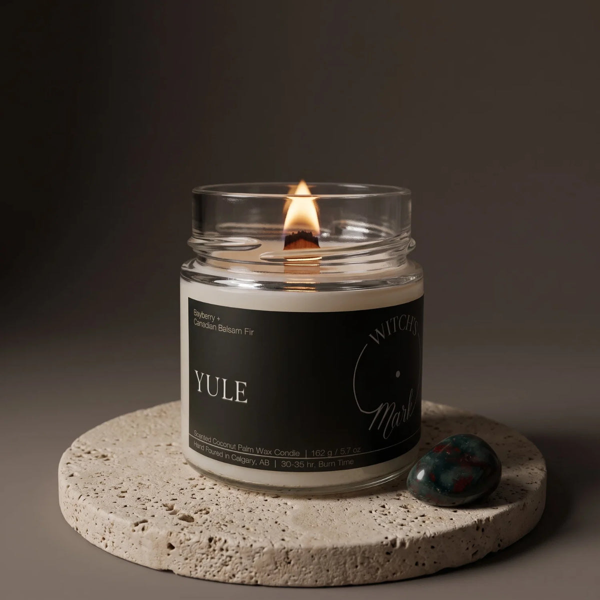 A glass jar candle called "Yule" is lit and sits on a porous stone coaster with a bloodstone crystal beside it.
