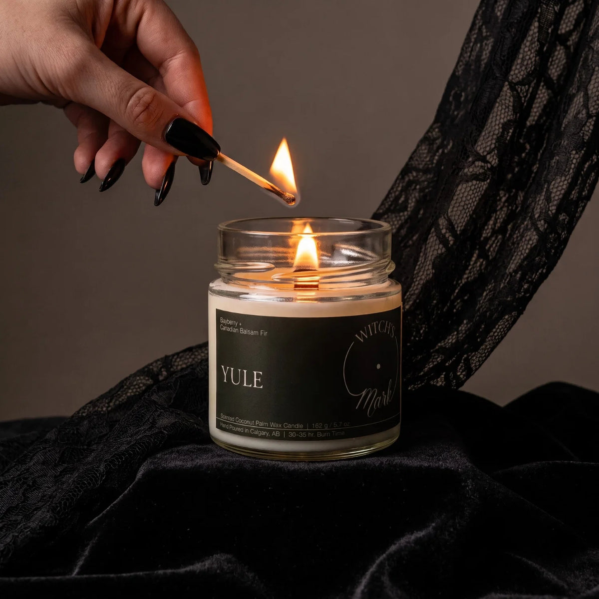 A witch's hand with black painted nails lights a candle called "Yule". The candle is sitting on top of black lace fabric.