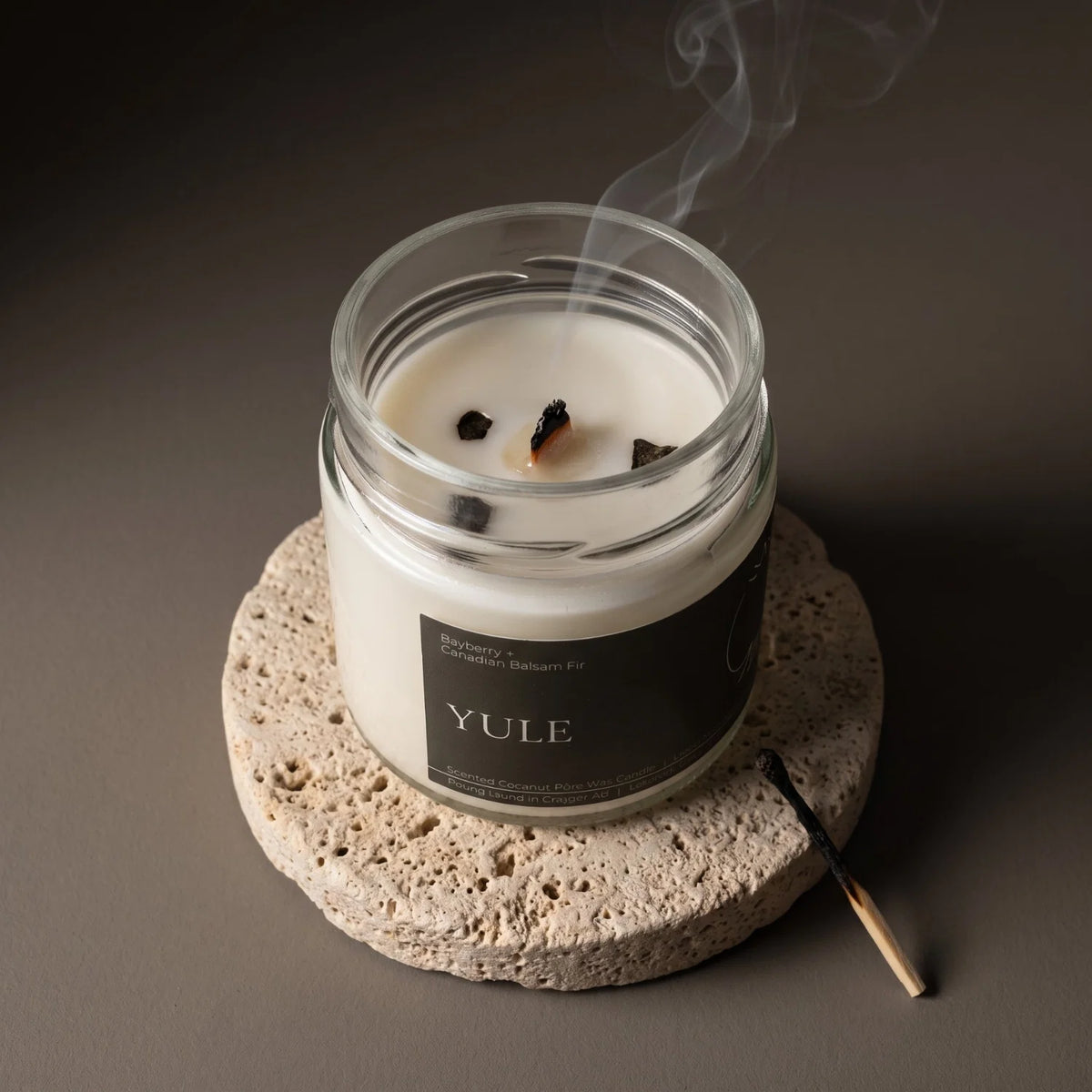 A coconut wax candle called "Yule" sits on a porous stone coaster with it's wick recently extinguished with swirls of white smoke.