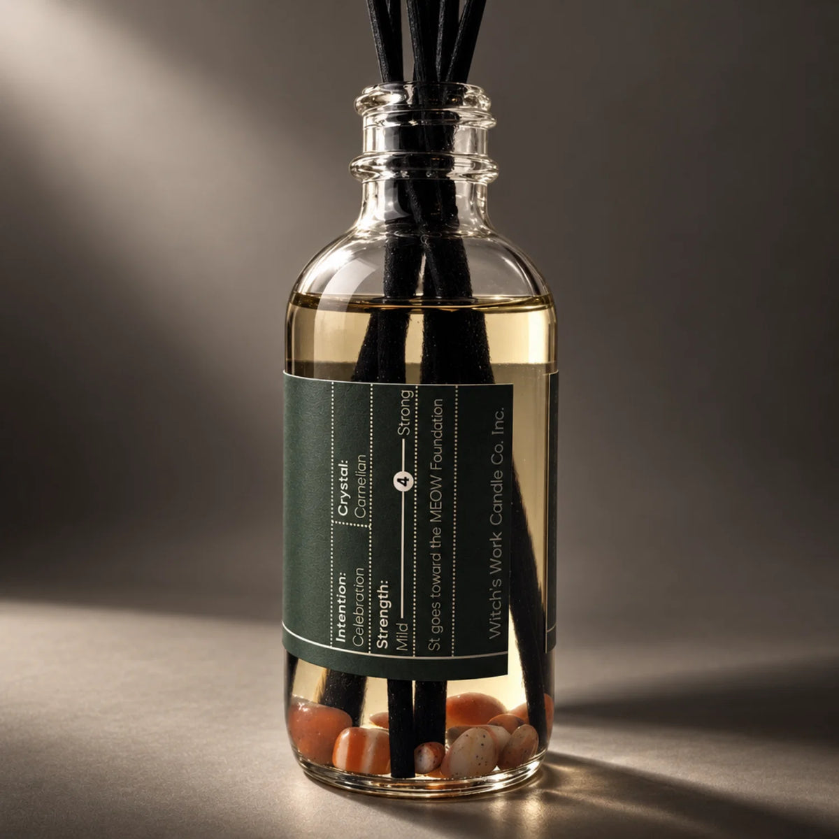 A Yule reed diffuser bottle with black reeds, a dark green label, and tiny carnelian crystals at the bottom, photographed on a dark gray studio background with dramatic side lighting.
