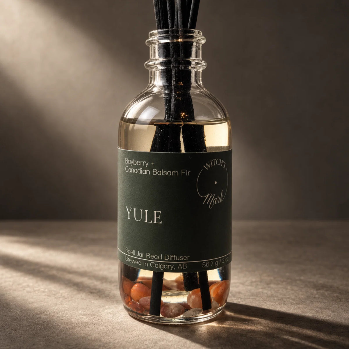 A glass reed diffuser bottle with a dark green label reading 'YULE' and 'Bayberry + Canadian Balsam Fir,' filled with clear liquid, black reeds, and tiny carnelian crystals, photographed on a warm brown surface with dramatic side lighting.