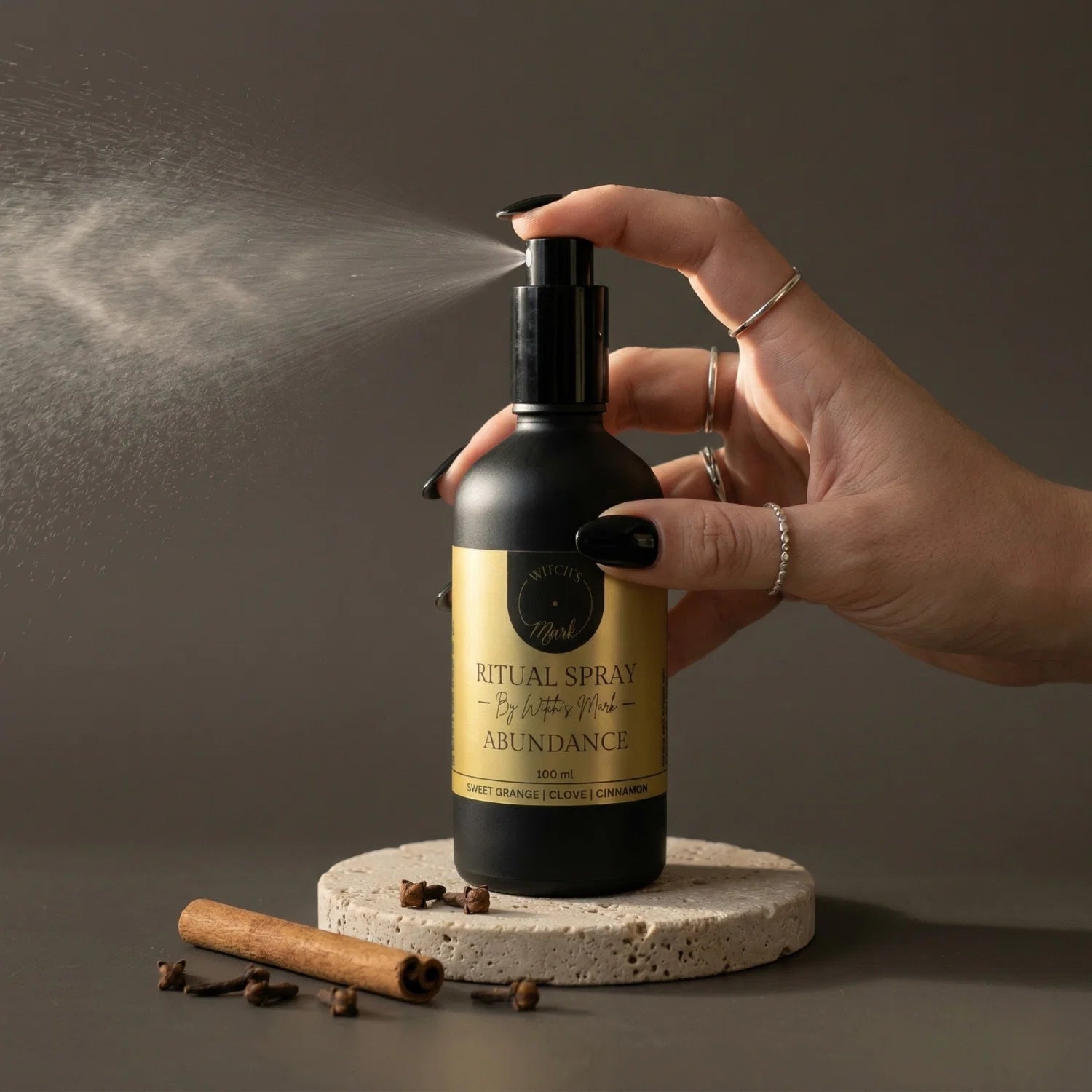 A hand with black nail polish spritzing the Witch's Mark Abundance ritual spray over cinnamon sticks and whole cloves. This prosperity aura mist is infused with citrine-charged moon water and warm essential oils to draw in limitless wealth and manifest bold intentions.