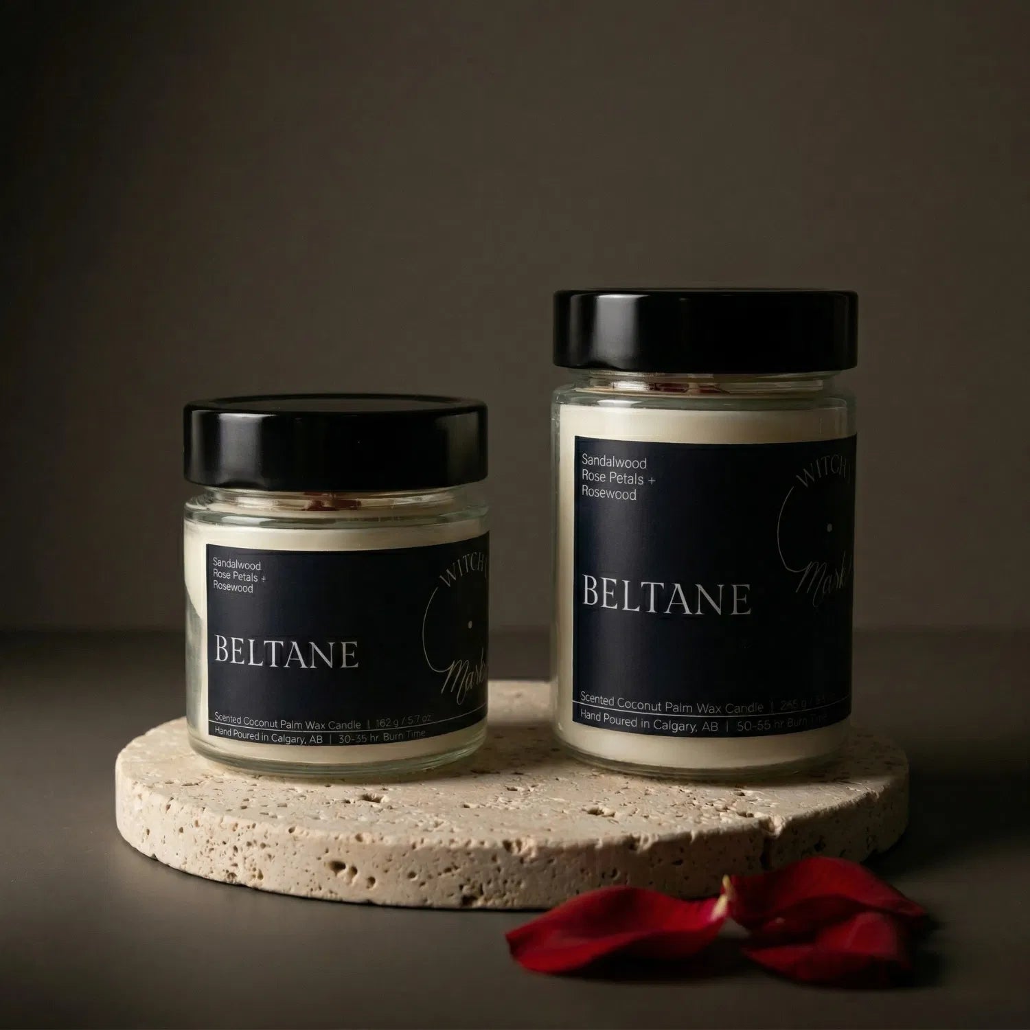 Two sizes of the seasonal Witch's Mark Beltane coconut wax candle, 5.7 oz and 8.6 oz, displayed on a round stone base and garnished with deep red rose petals.