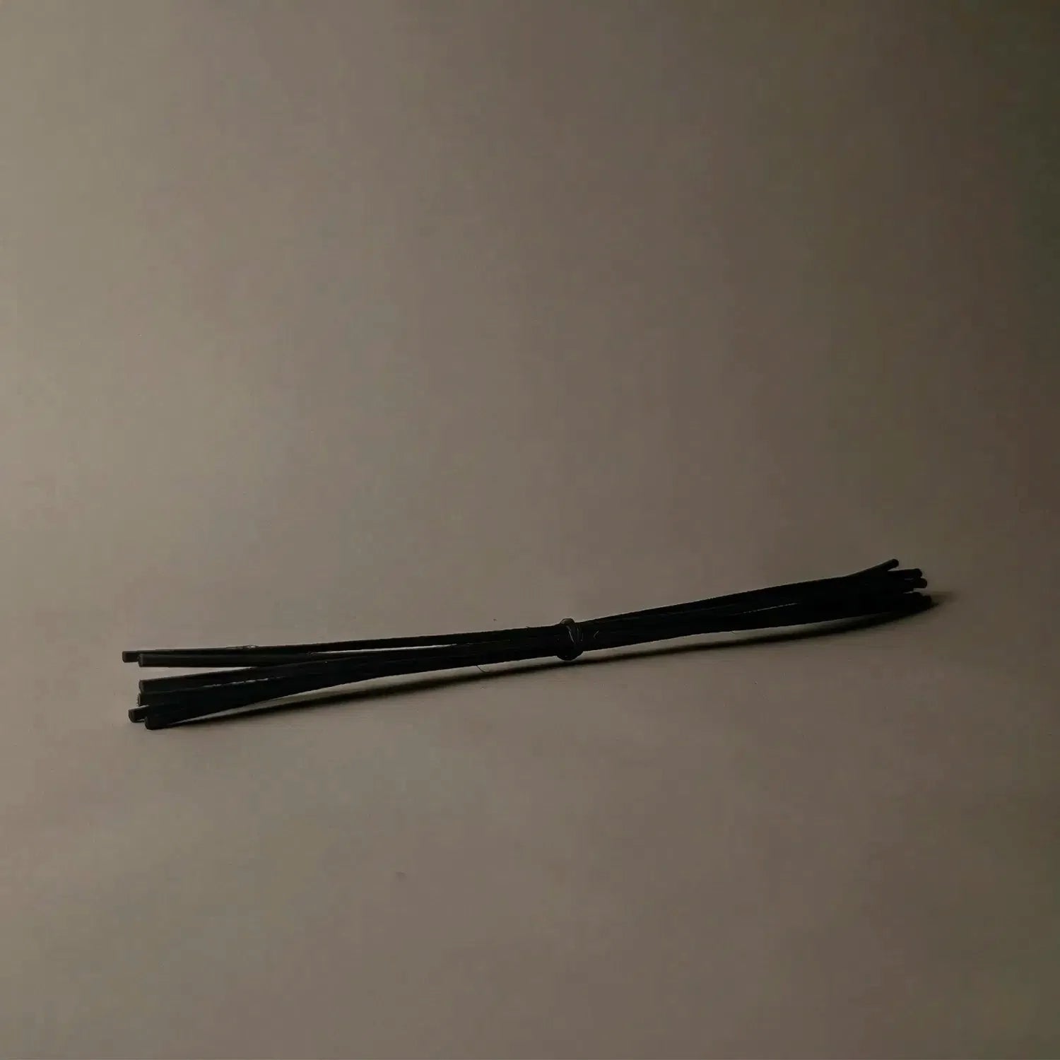 A convenient 6-pack bundle of sleek black rattan diffuser reeds resting horizontally against a dark background. Keeping a spare set of these replacement reeds ensures your space remains beautifully fragranced for weeks to come.