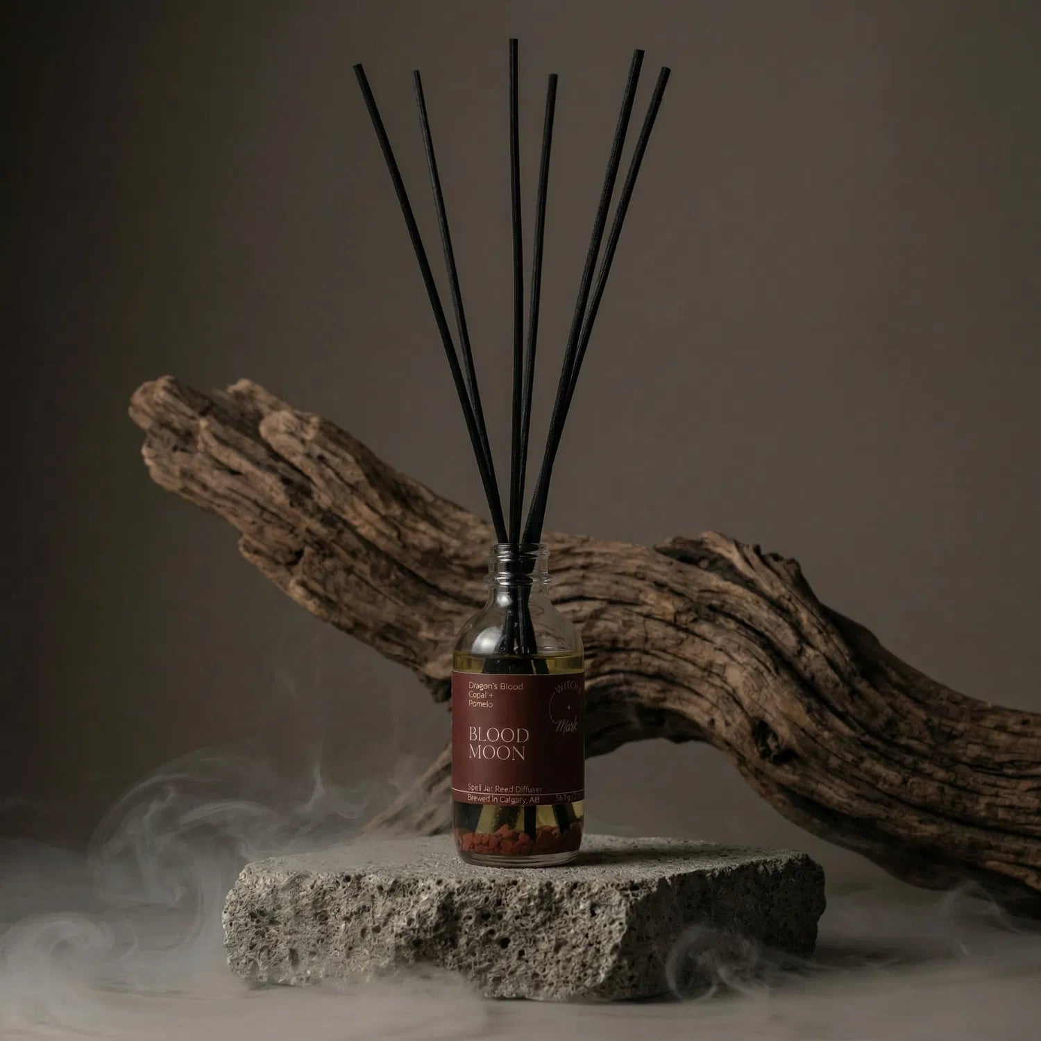 The Blood Moon apothecary spell jar reed diffuser by Witch's Mark sitting on a stone base, enveloped in atmospheric mist with natural driftwood in the background, perfectly setting the stage for raw manifesting power and lunar eclipse magick.