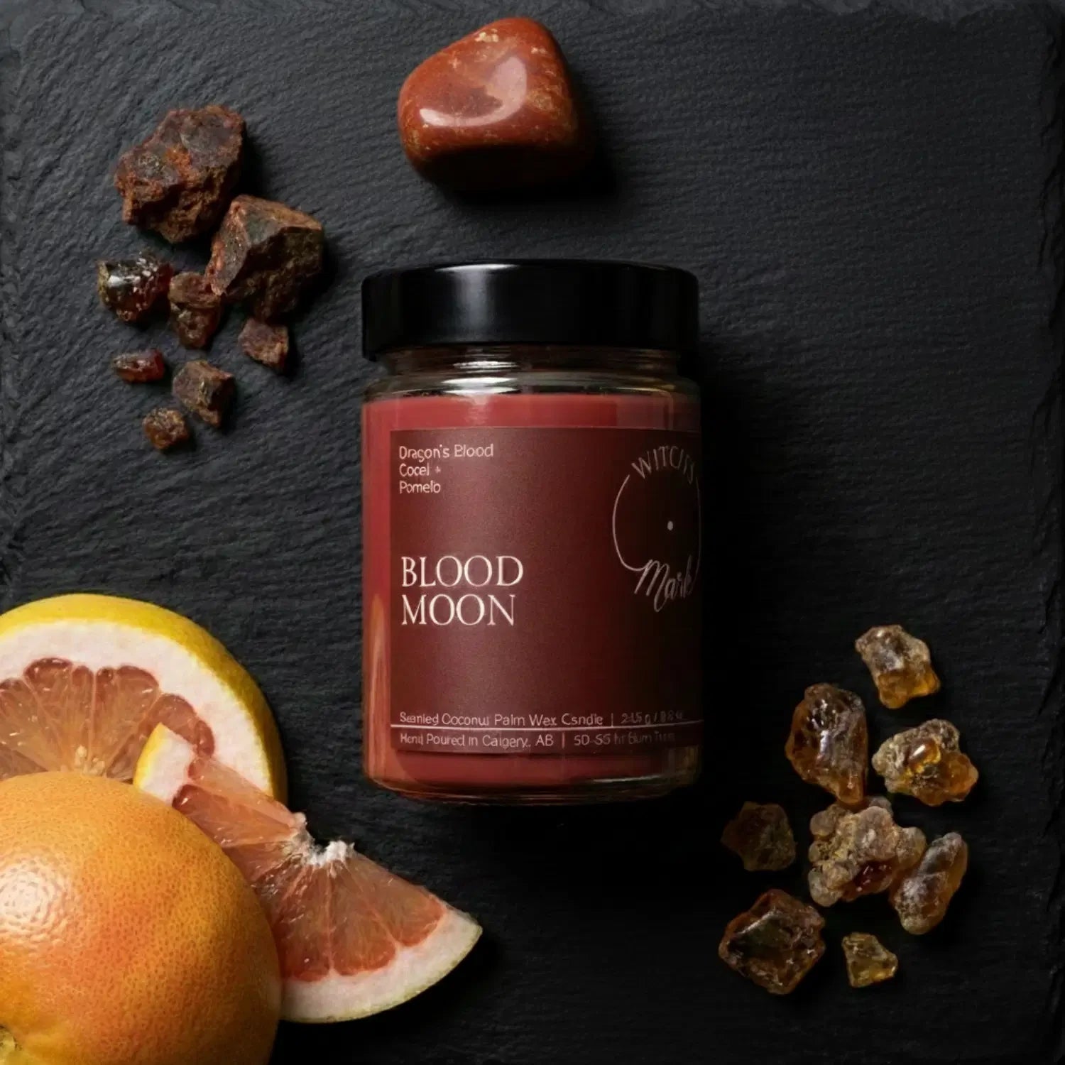 A top-down flat lay highlighting the invigorating scent profile of the Blood Moon candle, featuring the red wax jar surrounded by fresh pomelo citrus slices, sacred golden copal, and dark red dragon's blood resin on a dark slate background.