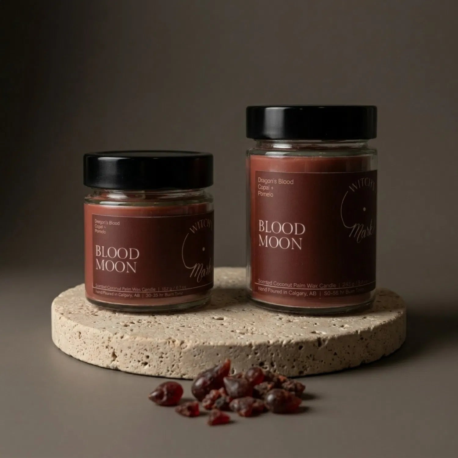 Two sizes of the Witch's Mark Blood Moon coconut wax candle, 5.7 oz and 8.6 oz, displaying the deep red wax on a round stone base, garnished with chunks of protective dragon's blood resin.