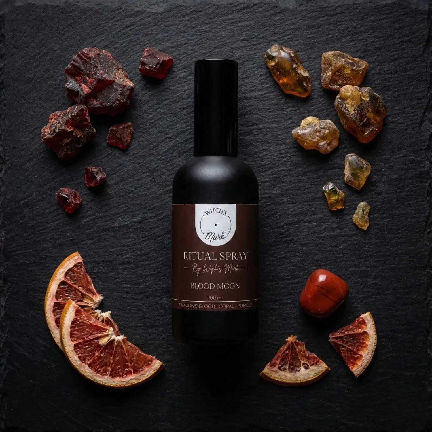 A top-down flat lay highlighting the empowering ingredients of the Blood Moon ritual mist by Witch's Mark. The black apothecary bottle sits surrounded by dark red chunks of ancient dragon's blood resin, golden pieces of sacred copal, and dried slices of bright pomelo against a dark slate background.