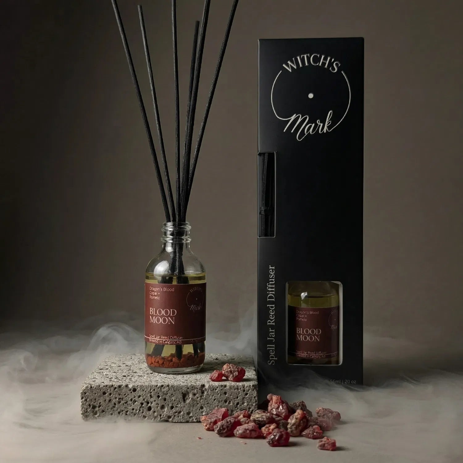 The Blood Moon spell jar reed diffuser by Witch's Mark displayed next to its sleek black packaging box. The apothecary glass vessel with black reeds rests on a textured stone block surrounded by mystical mist and scattered chunks of dark red dragon's blood resin, crafted to awaken elemental fire.
