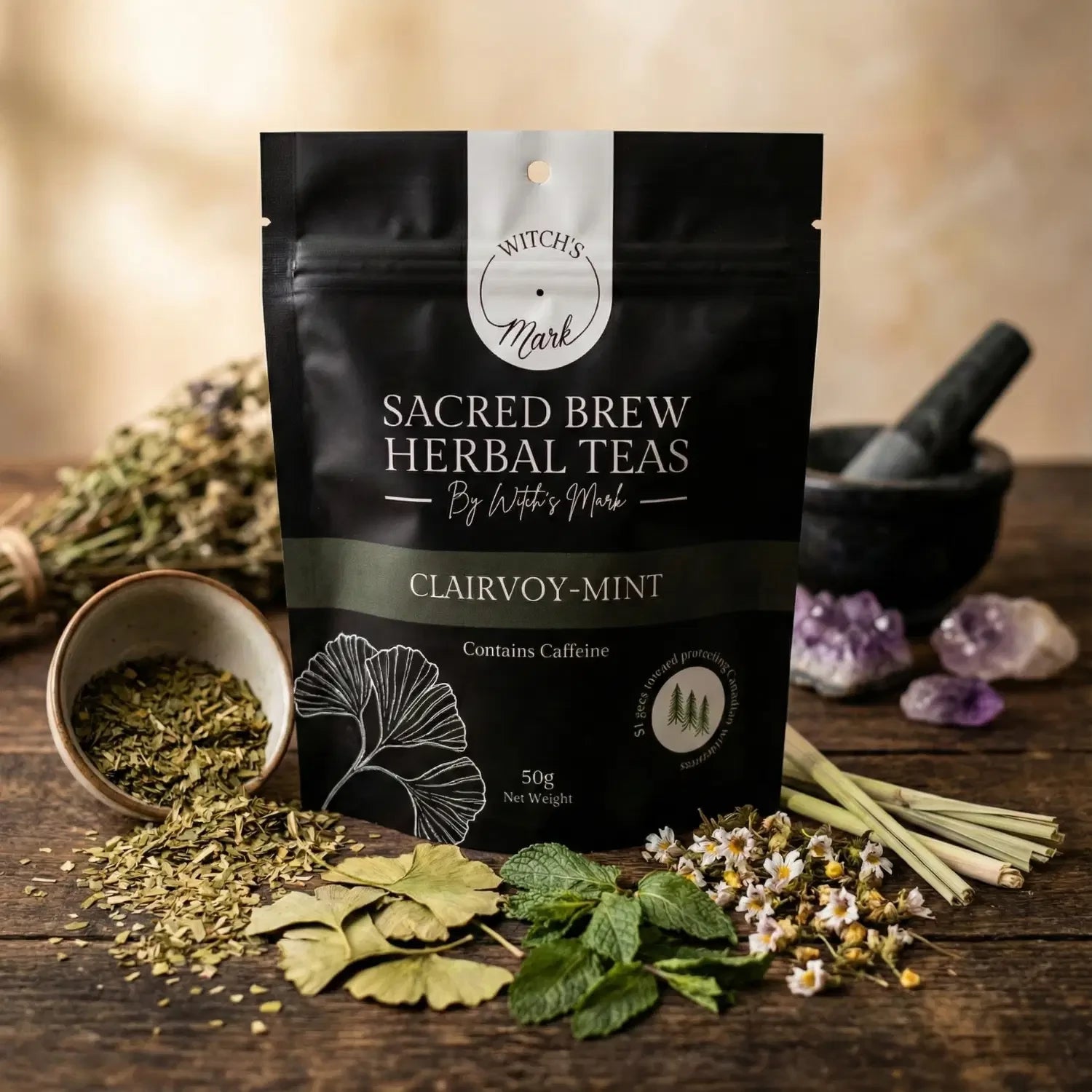 The Sacred Brew Herbal Teas Clairvoy-Mint pouch by Witch's Mark displayed on a rustic wooden table. This meditation tea blend is surrounded by its raw clarifying ingredients including yerba mate, ginkgo leaves, fresh peppermint, lemongrass, and eyebright, alongside amethyst crystals to help connect to your intuition.