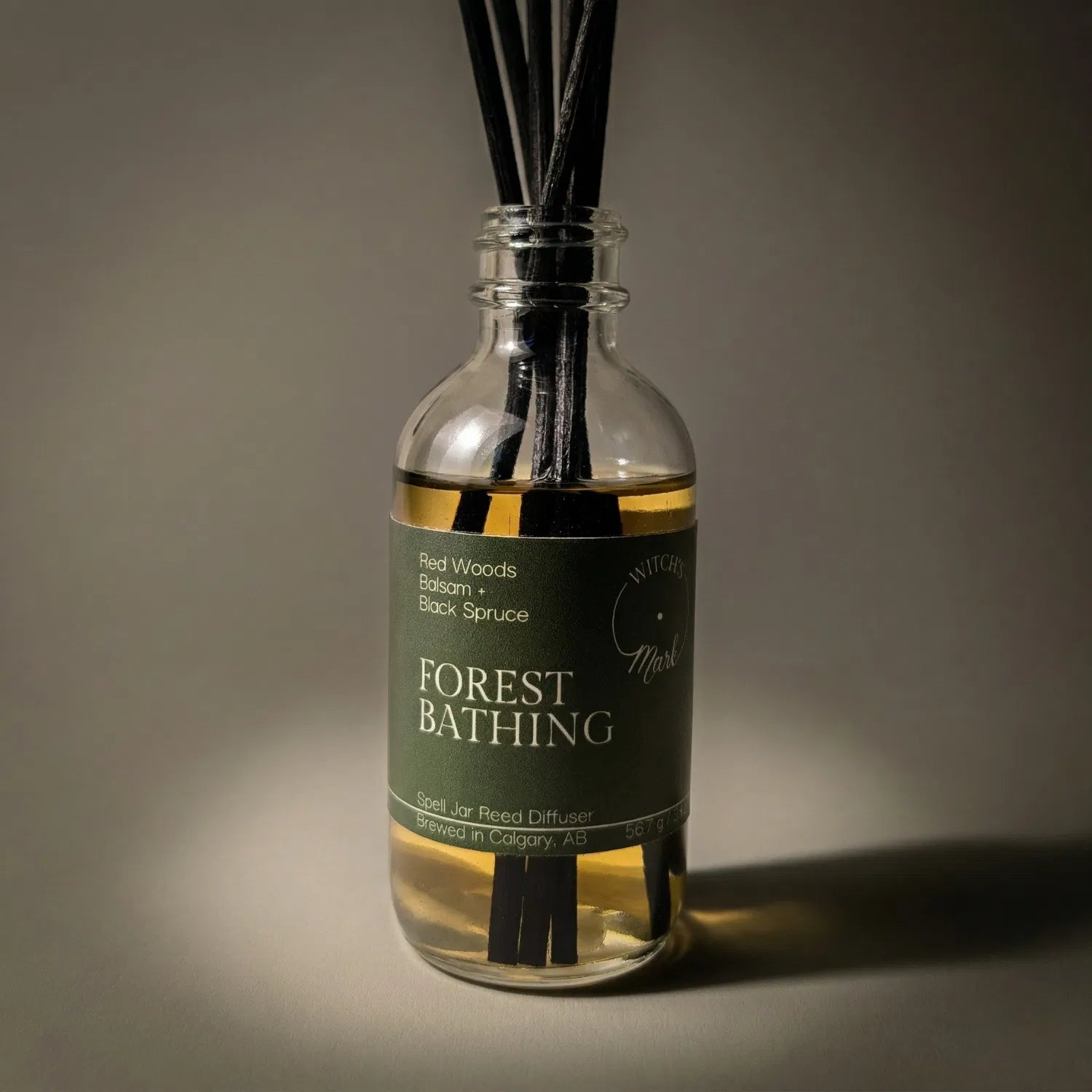 A dramatic spotlight close-up of the Forest Bathing spell jar reed diffuser, highlighting the deep green front label which lists its magical, grounding scent profile of ancient red woods, resinous balsam, and dark black spruce.