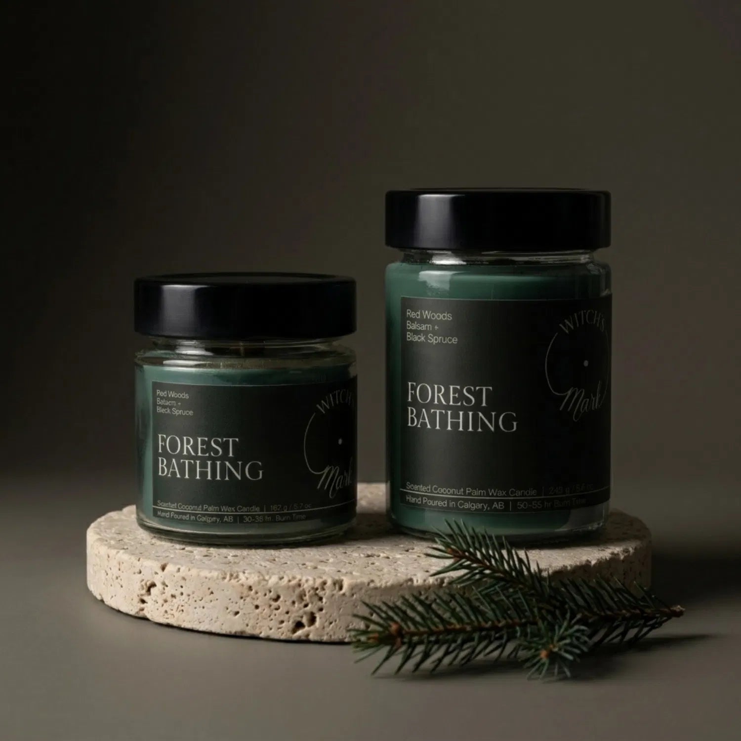 Two sizes of the Witch's Mark Forest Bathing coconut wax candle, 5.7 oz and 8.6 oz, displaying the deep green wax on a round stone base, garnished with a fresh sprig of black spruce.