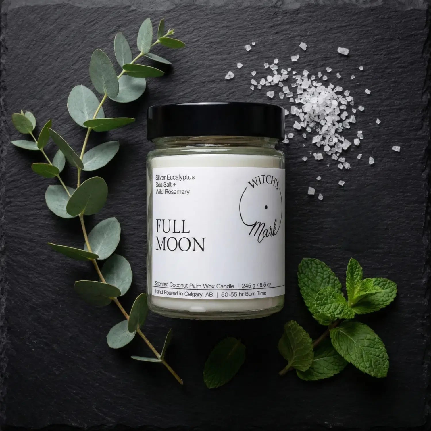 A top-down flat lay highlighting the cleansing ingredients of the Full Moon candle, featuring the jar surrounded by fresh silver eucalyptus leaves, sprigs of wild rosemary, and a scattering of coarse sea salt on a dark slate background.