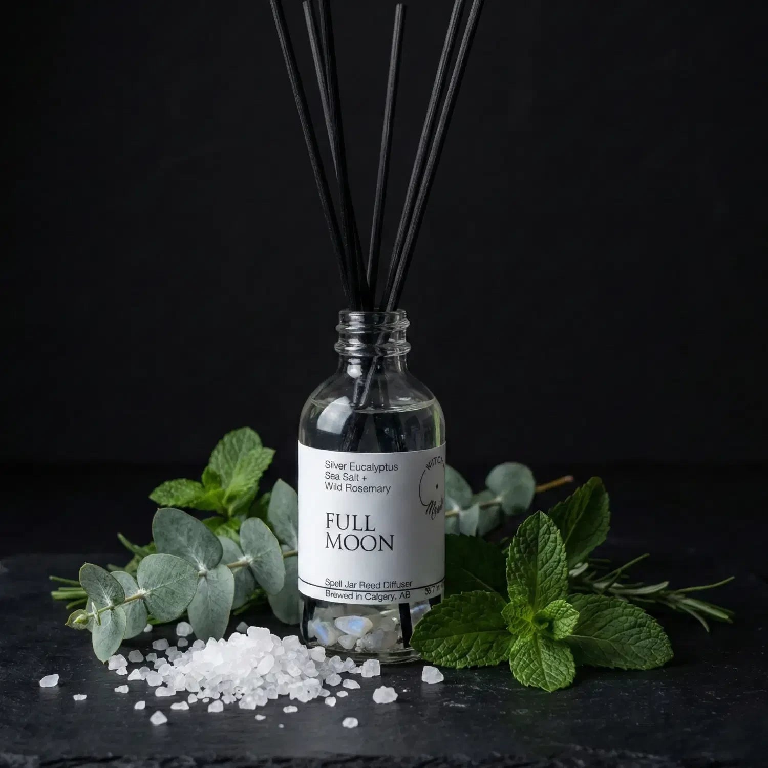 The Full Moon spell jar reed diffuser by Witch's Mark showcased against a dark slate background, surrounded by its core botanical ingredients: fresh silver eucalyptus branches, sprigs of wild rosemary, and a scattering of purifying coarse sea salt.
