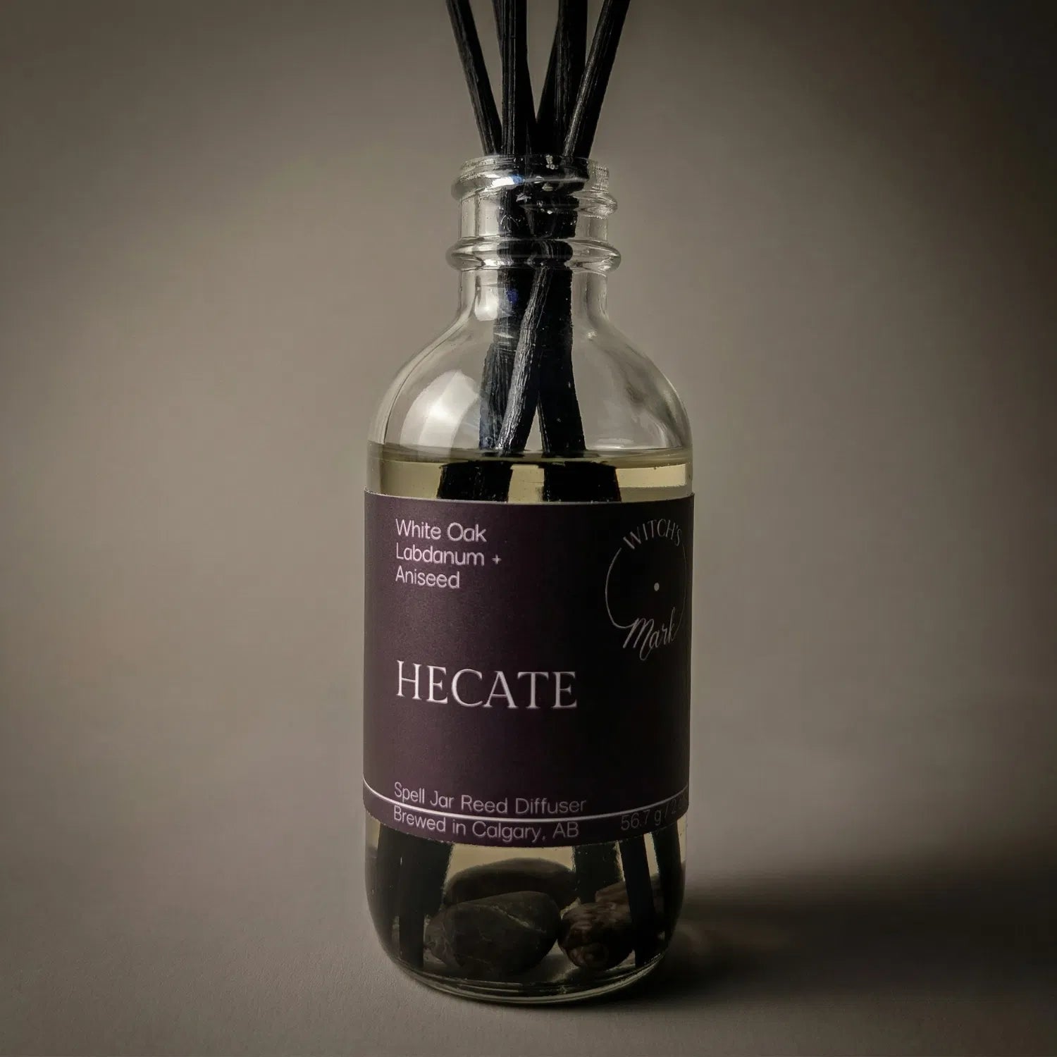 A dramatic spotlight close-up of the Hecate spell jar reed diffuser, highlighting the deep purple front label which lists its magical, ancient scent profile of grounded white oak, resinous labdanum, and spellbinding aniseed.