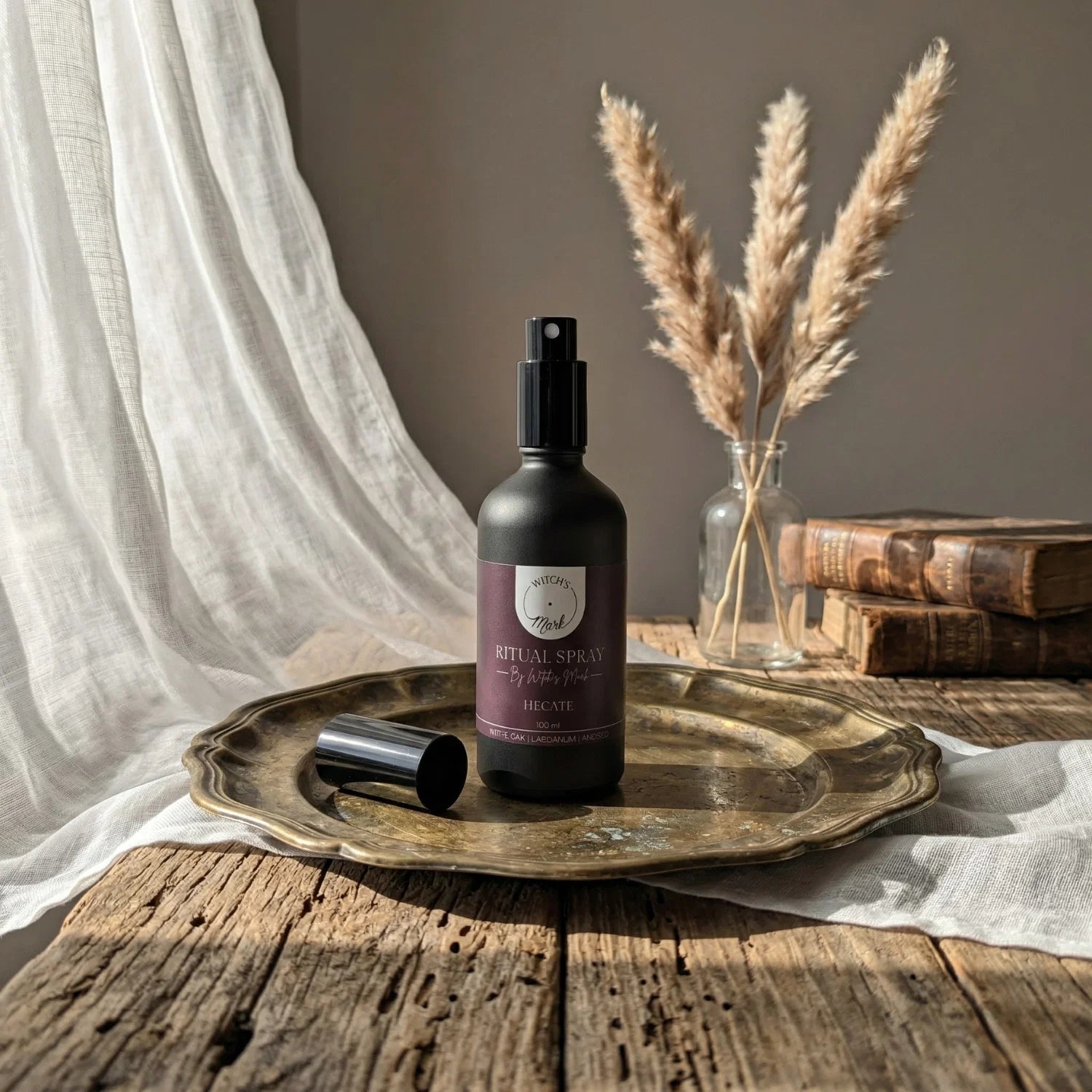The matte black bottle of Hecate ritual spray with its dark purple label elegantly displayed on a vintage brass tray atop a rustic wooden table bathed in soft light. This sacred elixir is perfect for misting over your altar to empower powerful shadow work and illuminate your spiritual journey at the crossroads.