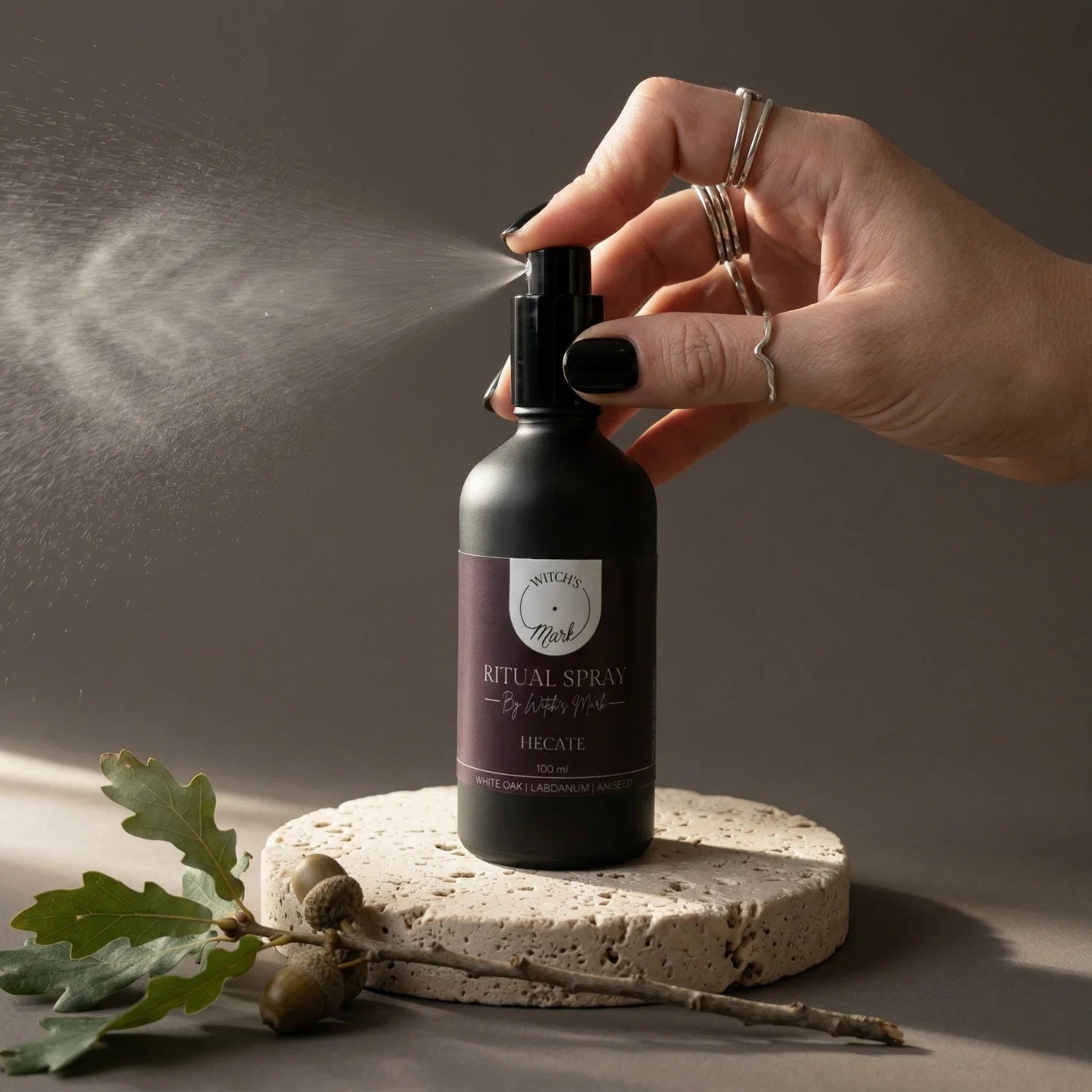 A hand with black nail polish spritzing the Witch's Mark Hecate ritual spray over a grounding sprig of white oak leaves and acorns. This transformative aura mist is infused with labradorite charged moon water to invoke the magick of the liminal realm and guide profound metamorphosis.