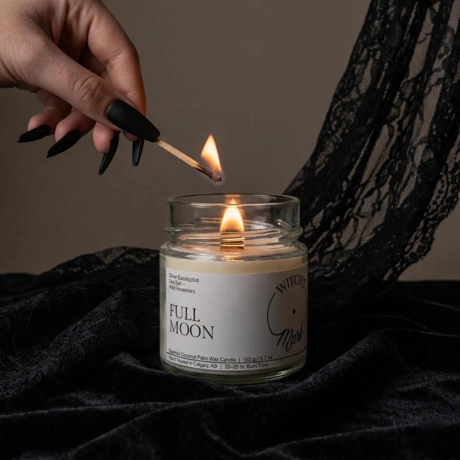 A hand with black nail polish using a match to light the wooden wick of a Full Moon intention candle by Witch's Mark, set against a dark aesthetic background featuring draped black lace.