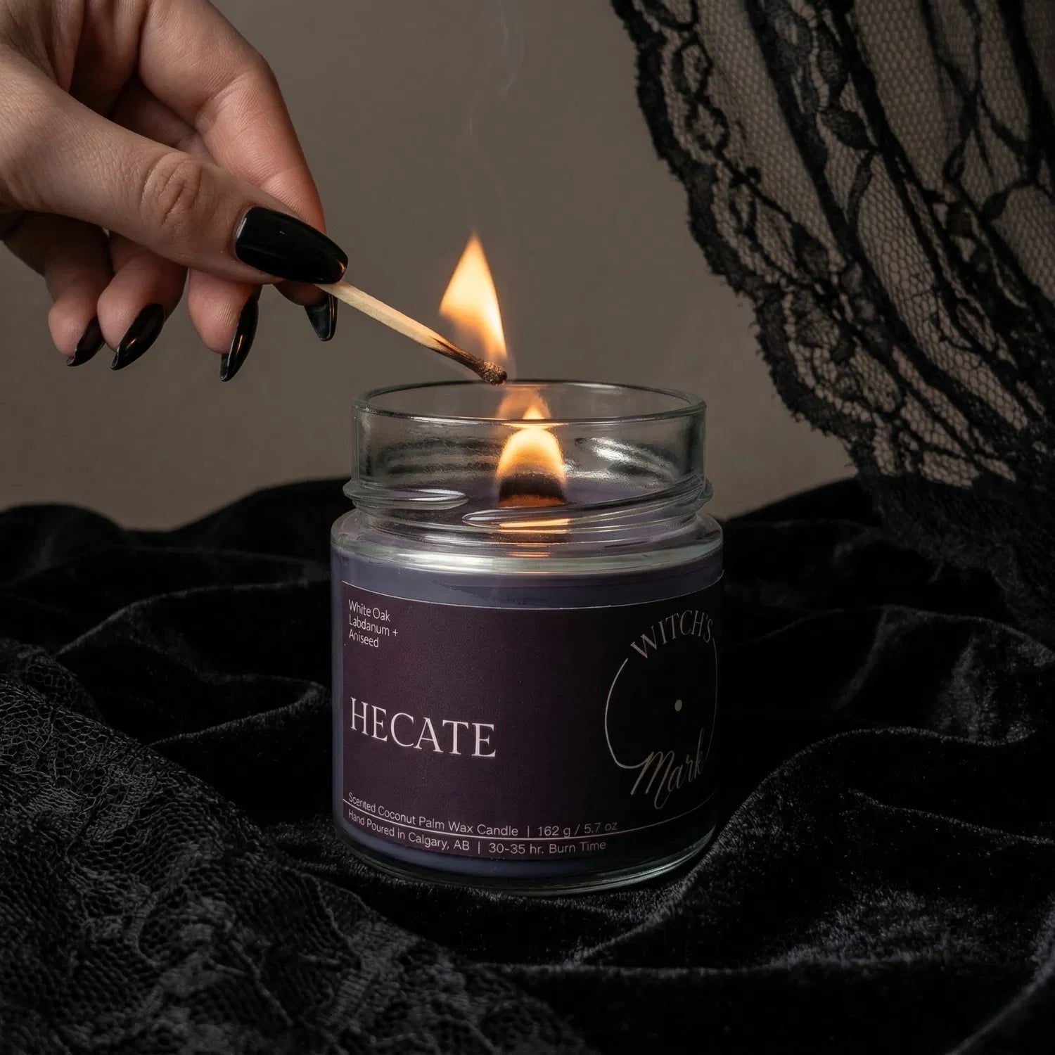 A hand with black nail polish using a match to light the wooden wick of a deep purple Hecate magic candle by Witch's Mark, set against a dark aesthetic background featuring draped black lace.