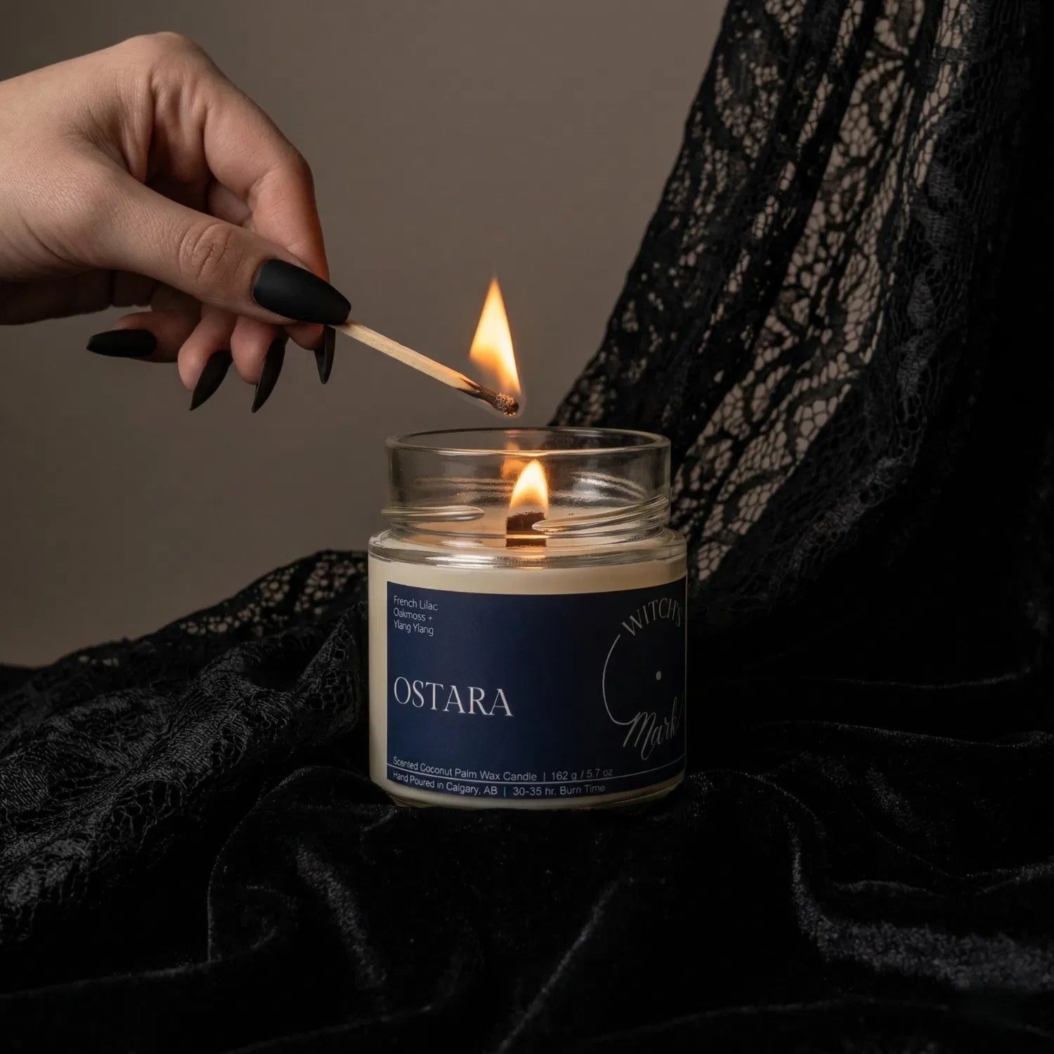 A hand with black nail polish using a match to light the wooden wick of an Ostara spring equinox candle by Witch's Mark, set against a dark aesthetic background featuring draped black lace.