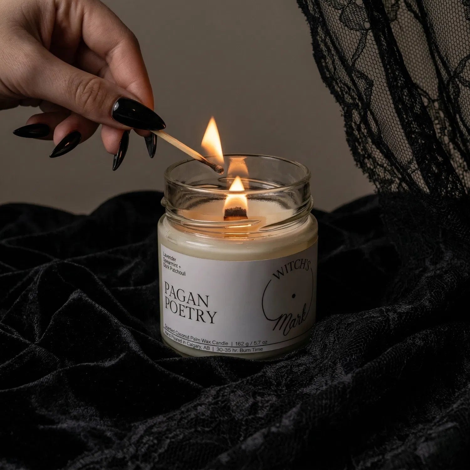 A hand with black nail polish using a match to light the wooden wick of a Pagan Poetry crystal candle by Witch's Mark, set against a dark aesthetic background featuring draped black lace.