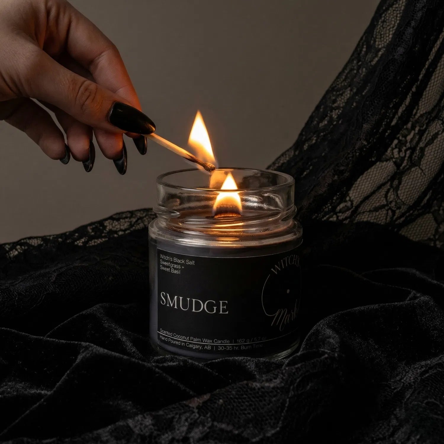 A hand with black nail polish using a match to light a Smudge wooden wick candle, set against a dark aesthetic background featuring draped black lace.