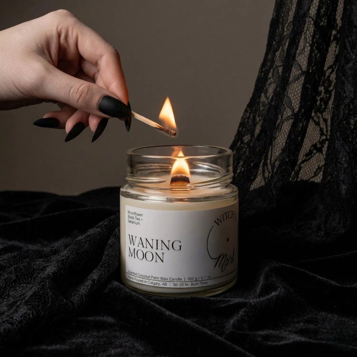 A hand with black nail polish using a match to light the wooden wick of a Waning Moon intention candle by Witch's Mark, set against a dark aesthetic background featuring draped black lace.