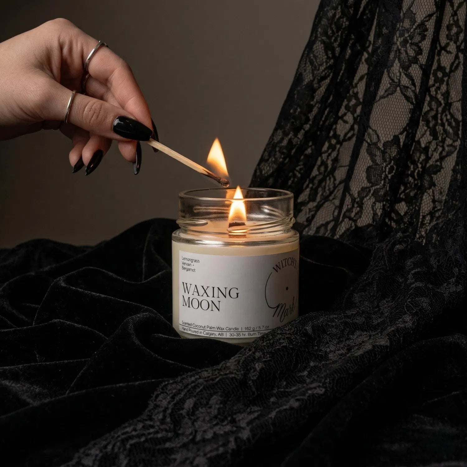 A hand with black nail polish using a match to light the wooden wick of a Waxing Moon intention candle by Witch's Mark, set against a dark aesthetic background featuring draped black lace.