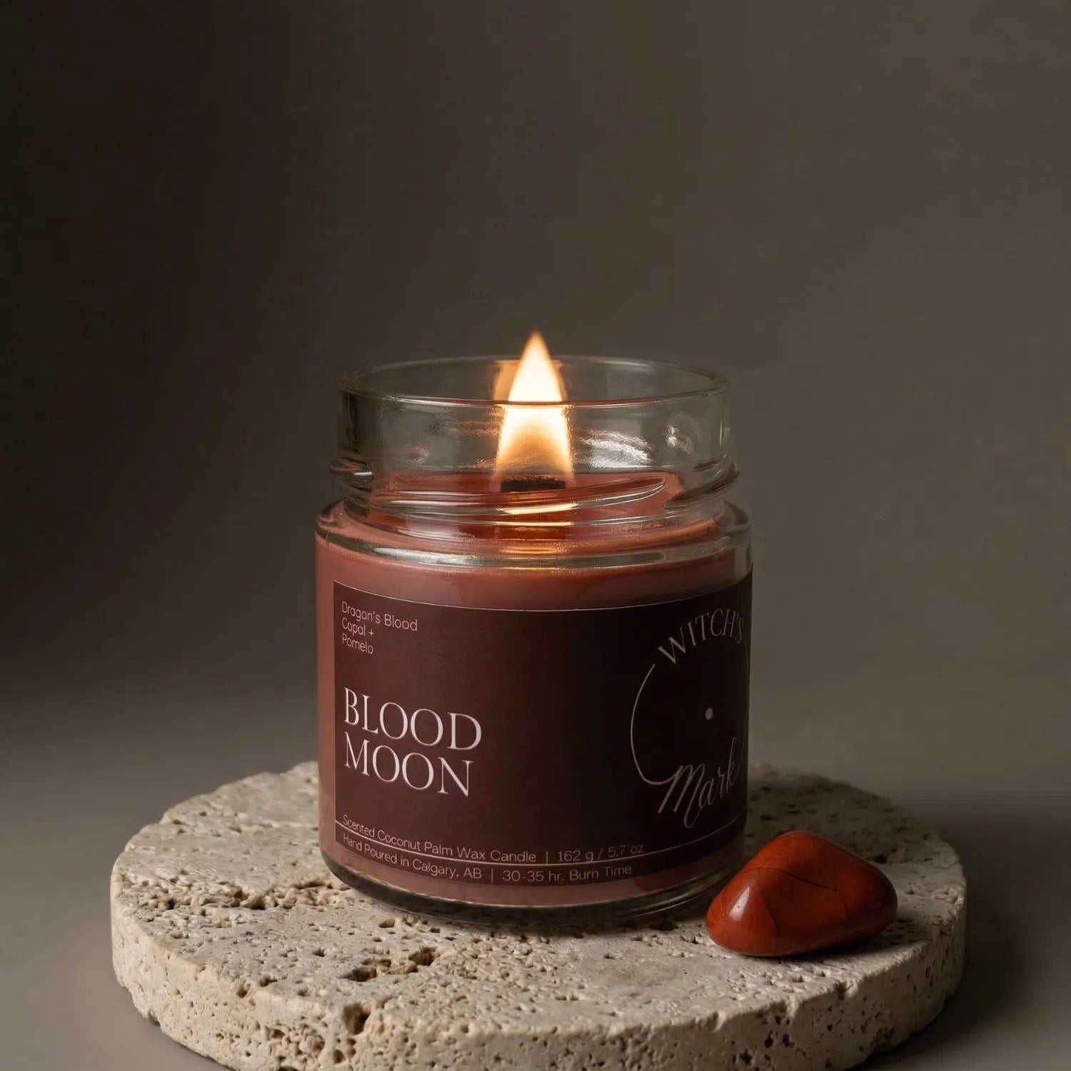A lit Blood Moon crystal candle by Witch's Mark resting on a textured stone coaster next to a grounding red jasper crystal. The deep red wax is designed to promote strength, courage, and protection during spiritual journeys.