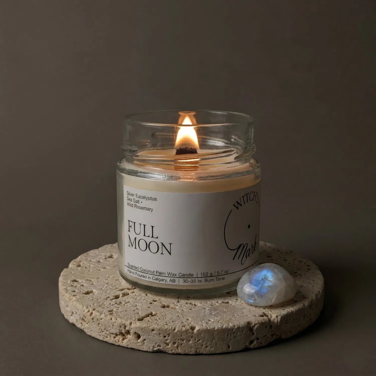 A lit Full Moon crystal candle by Witch's Mark resting on a textured stone coaster next to a shimmering moonstone crystal, crafted to enhance intuition and emotional balance.