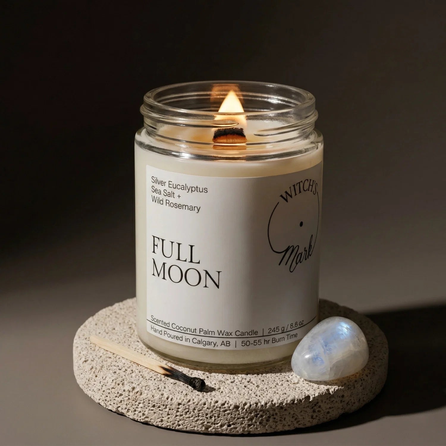 A lit Full Moon candle from the Lunar Cycle bundle resting on a stone coaster, accompanied by a glowing moonstone crystal designed to enhance intuition and step into personal power.