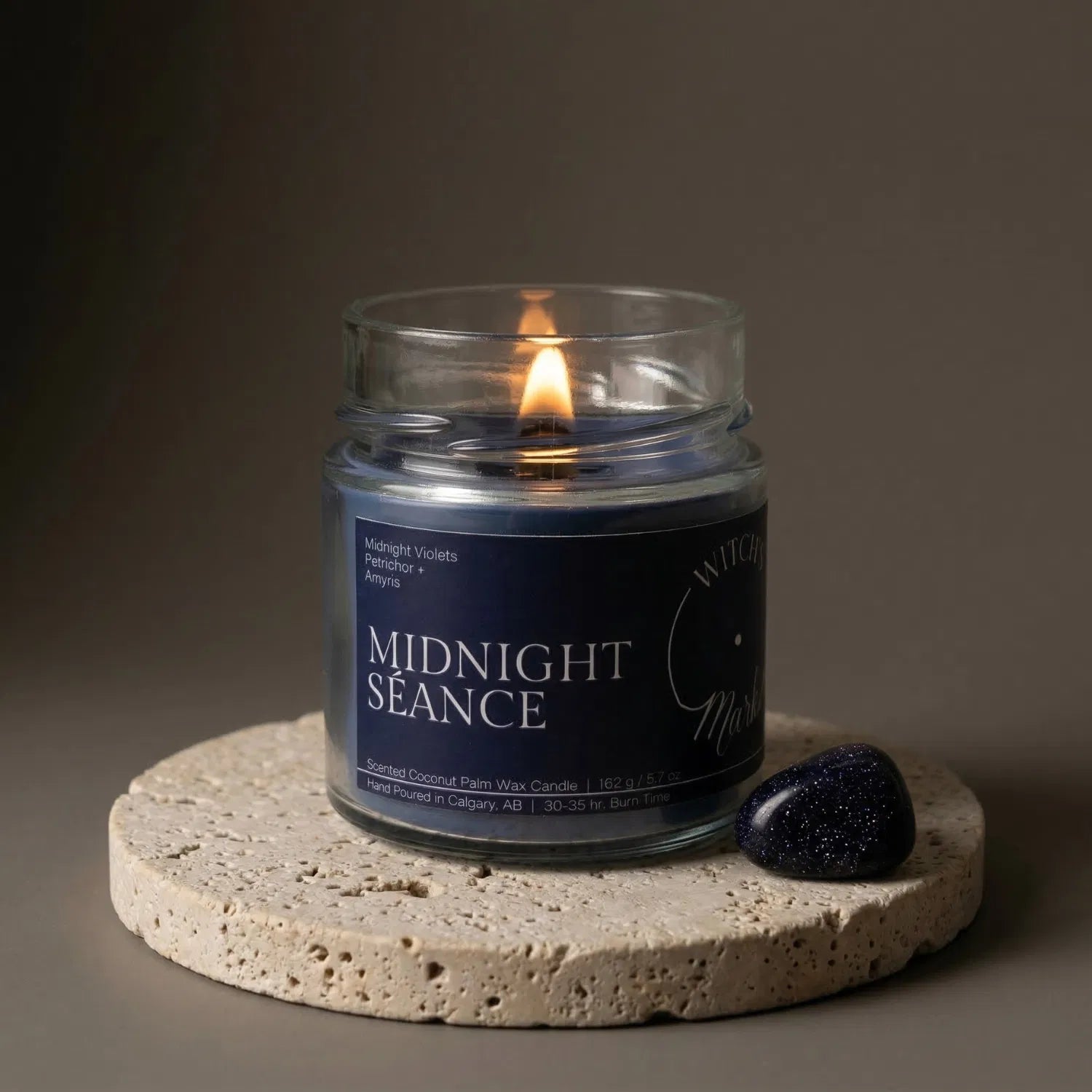 A lit Midnight Séance crystal candle by Witch's Mark resting on a textured stone coaster next to a grounding tree agate crystal. The deep blue wax is designed to promote throat chakra communication and clarity during meditative journeys.