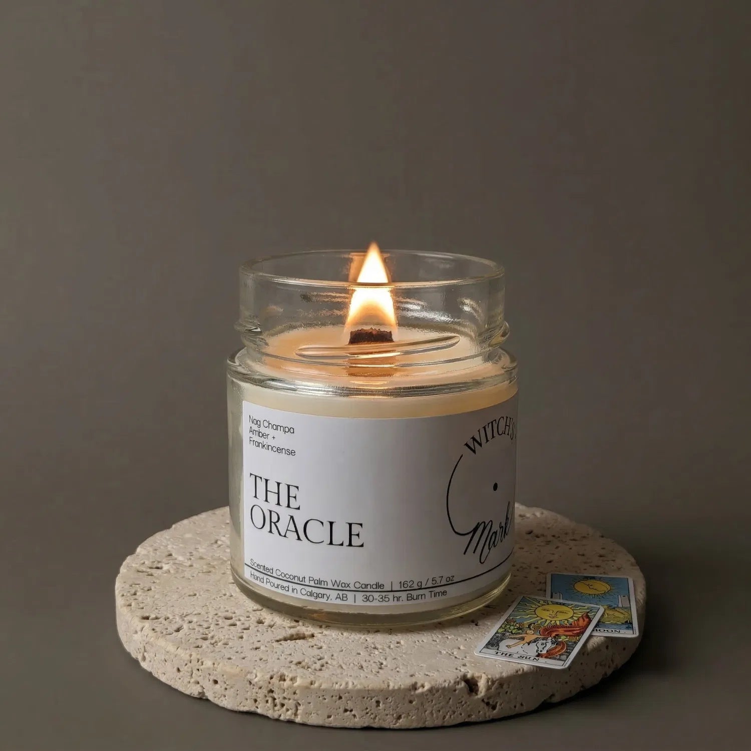 A lit Oracle tarot candle by Witch's Mark resting on a textured stone coaster alongside two mini tarot cards, representing the hidden reading waiting at the bottom of the jar.