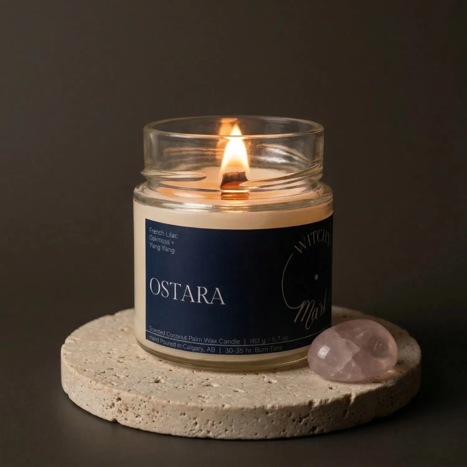 A lit Ostara crystal candle by Witch's Mark resting on a textured stone coaster next to a smooth pink rose quartz crystal, crafted to encourage self-love, harmony, and spring renewal.