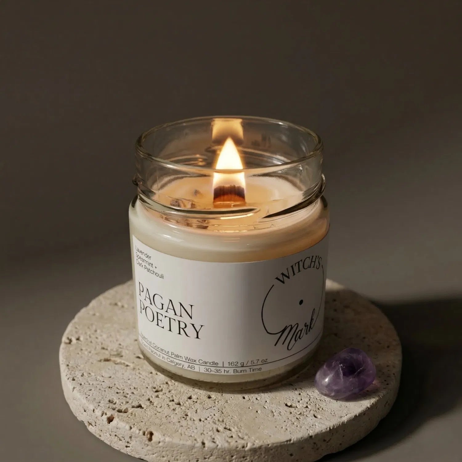 A gently lit Pagan Poetry candle sitting on a textured stone coaster next to a purple amethyst crystal, highlighting the tranquil ambiance and stress-relieving properties of the lavender and patchouli blend.