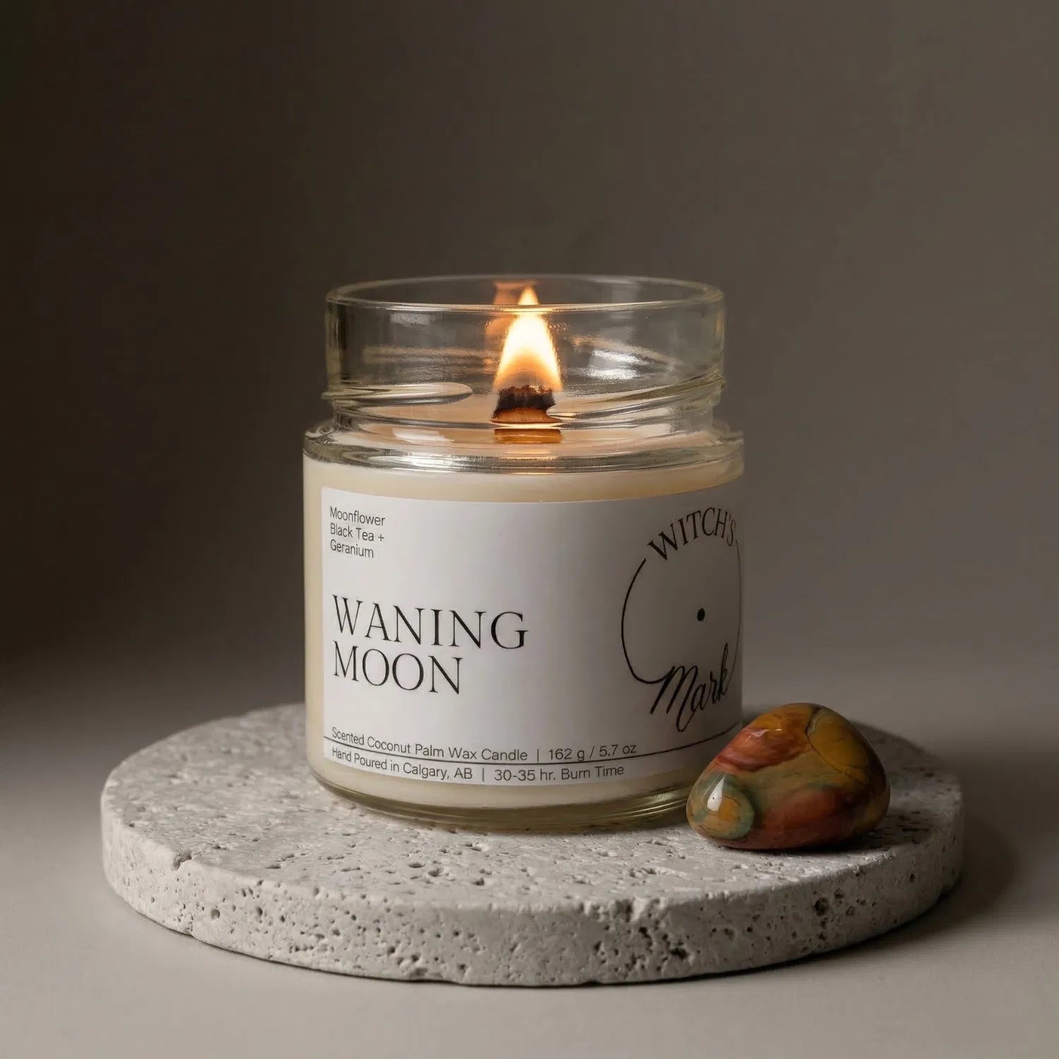 A lit Waning Moon crystal candle by Witch's Mark resting on a textured stone coaster next to a smooth polychrome jasper stone, crafted to encourage calming energy and introspection.